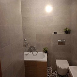 Piotr Wuttke I Andrzej Lotka - Wykończona łazienka z wanną obudowaną drewnem, toaletą, umywalką i czarno-białą wzorzystą podłogą, szare płytki na ścianach, dekoracyjne rośliny doniczkowe.