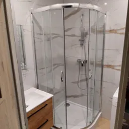 Nowoczesna łazienka z kabiną prysznicową narożną, umywalką w szafce z drewnianymi frontami i marmurowymi płytkami na ścianach, widok zza framugi drzwi.