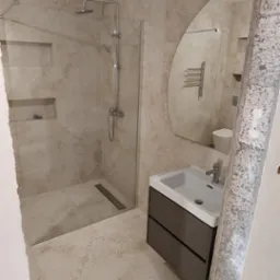 Nowoczesna łazienka w beżowych płytkach z kabiną prysznicową typu walk-in, umywalką z szafką i asymetrycznym lustrem, widoczny surowy betonowy fragment framugi drzwi.