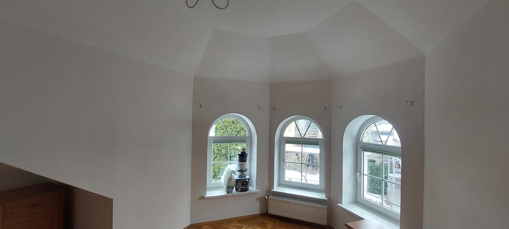 Pomalowane na biało wnętrze z trzema łukowatymi oknami, na parapecie jednego z nich ustawione wiaderka z farbą, parkiet na podłodze.