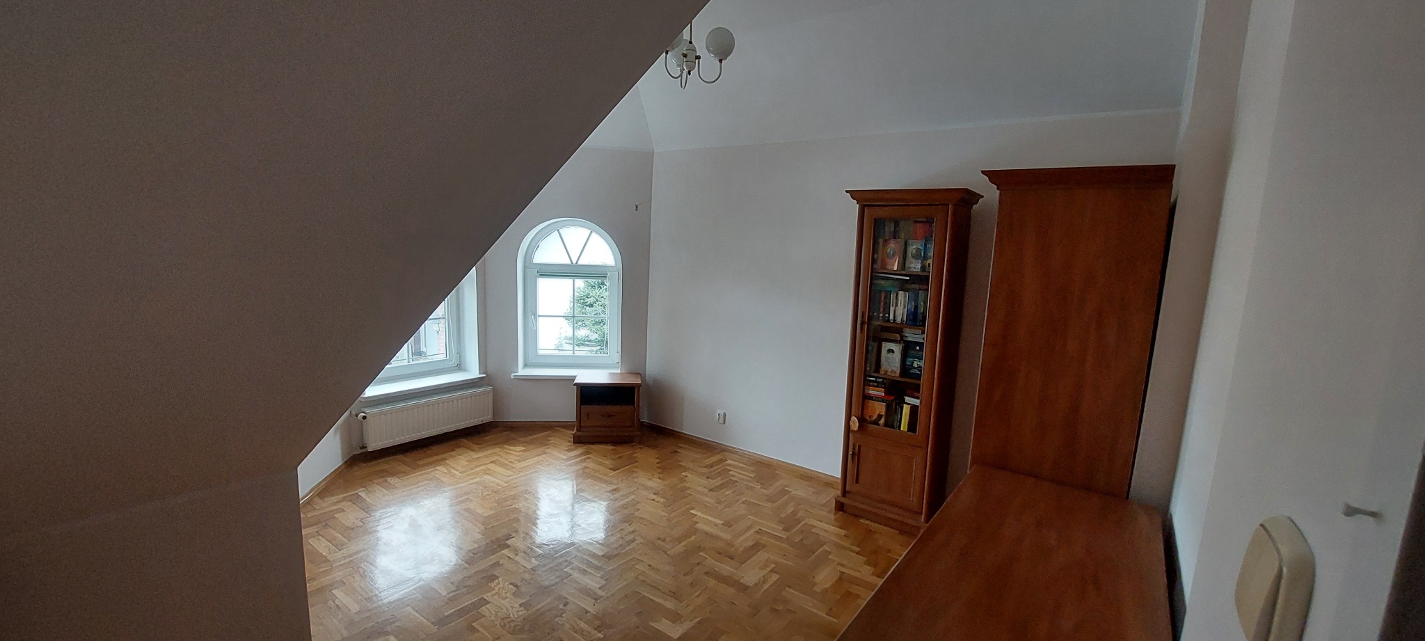 Jasne wnętrze poddasza z parkietem w jodełkę, oknami w kształcie łuku i drewnianymi meblami, widok z narożnika pokoju.