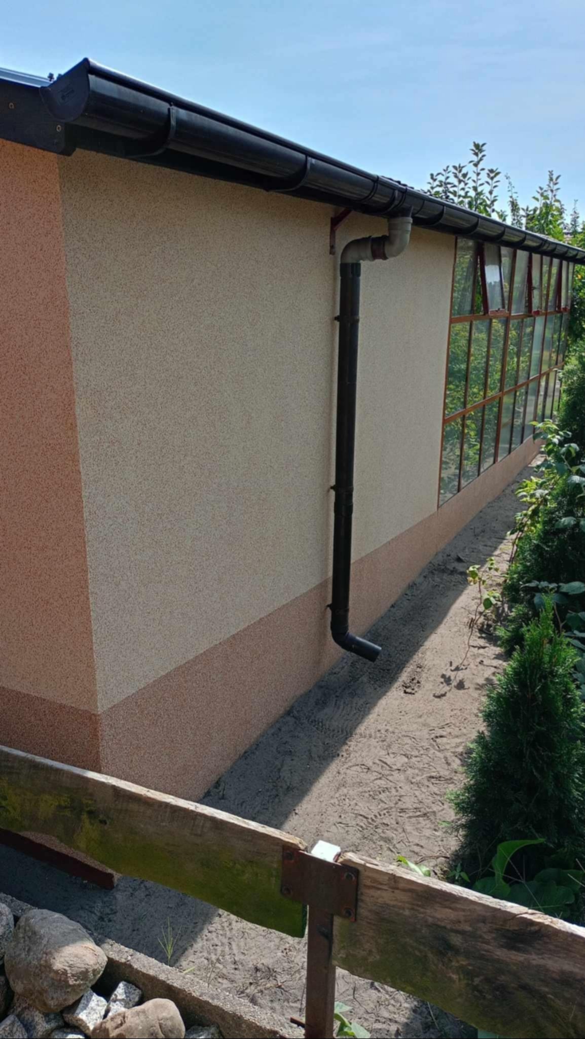Elewacja budynku z oknami szklarniowymi, dwukolorowym tynkiem i czarną rynną spustową, widok z zewnątrz.