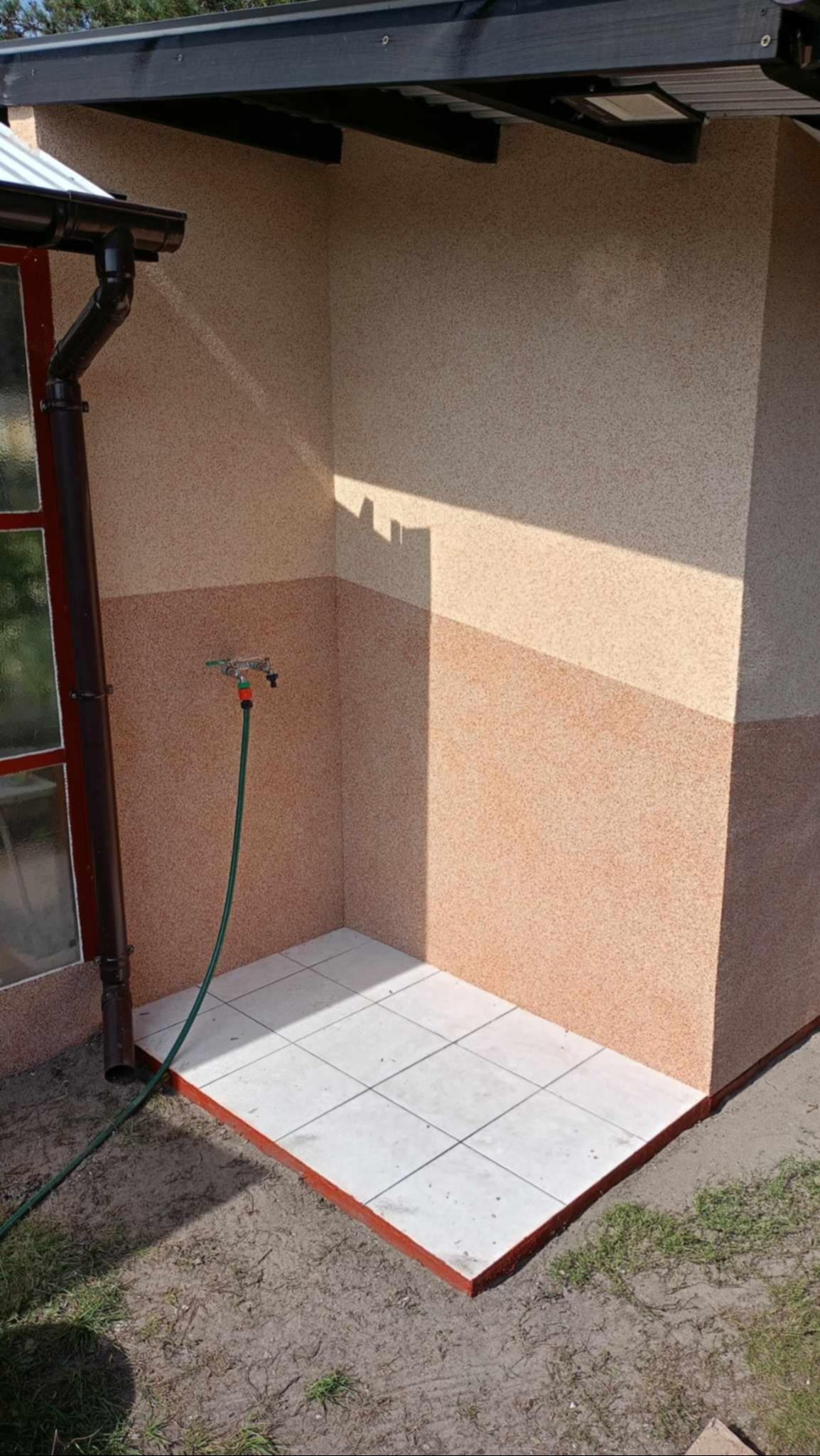 Zewnętrzny prysznic z wyłożoną płytkami podłogą, dwukolorową elewacją i zielonym wężem ogrodowym.