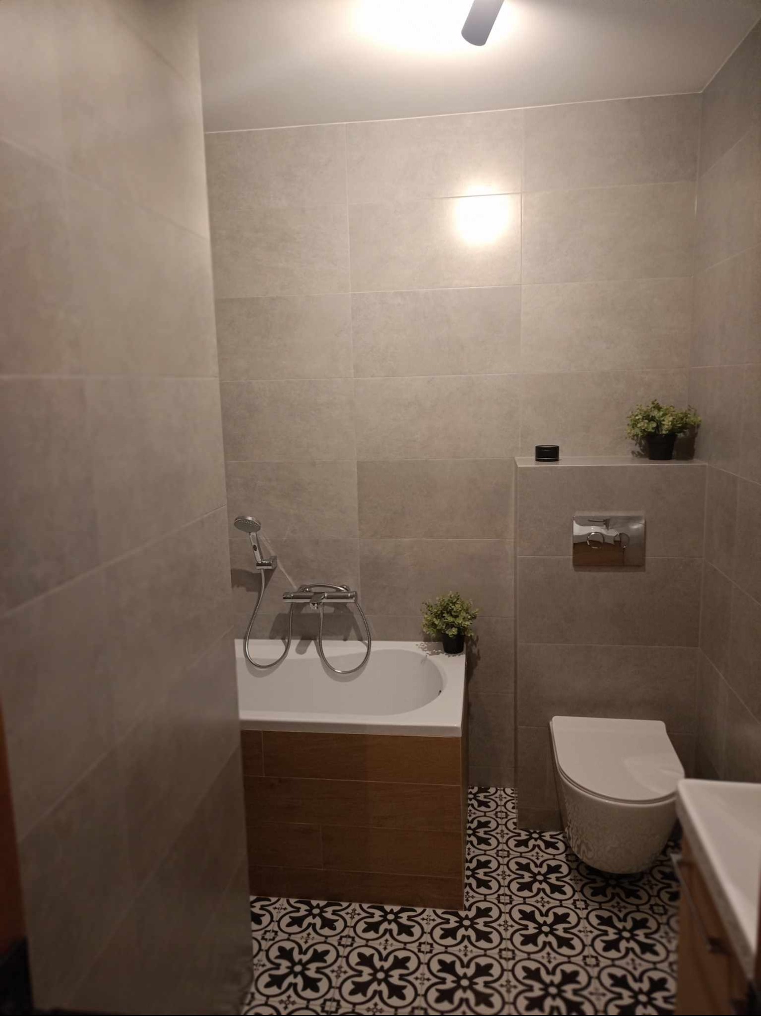 Wykończona łazienka z wanną obudowaną drewnem, toaletą, umywalką i czarno-białą wzorzystą podłogą, szare płytki na ścianach, dekoracyjne rośliny doniczkowe.