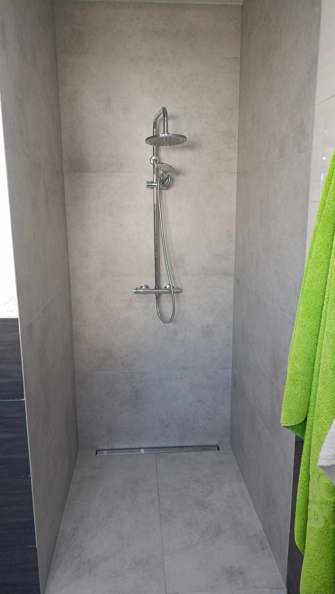 Nowoczesny prysznic z szarymi płytkami imitującymi beton, stalowa deszczownica i odpływ liniowy, z zielonym ręcznikiem po prawej stronie.