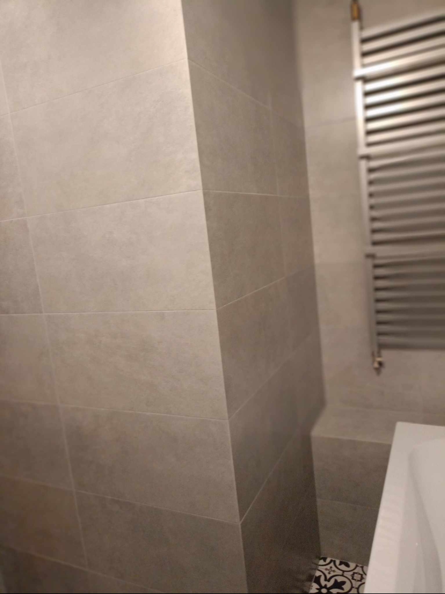 Jasnoszare, matowe płytki ceramiczne ułożone poziomo na ścianie łazienki, widoczny fragment grzejnika drabinkowego i wanny, dekoracyjne płytki podłogowe w rogu.