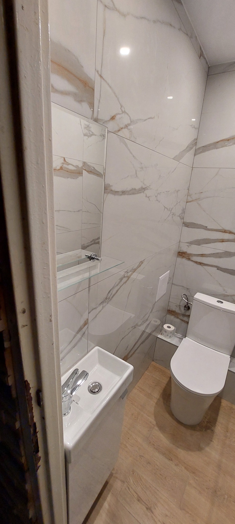 Wykończona łazienka z białymi płytkami imitującymi marmur, mała umywalka z szafką, toaleta i drewnopodobna podłoga, widok zza uchylonych drzwi.