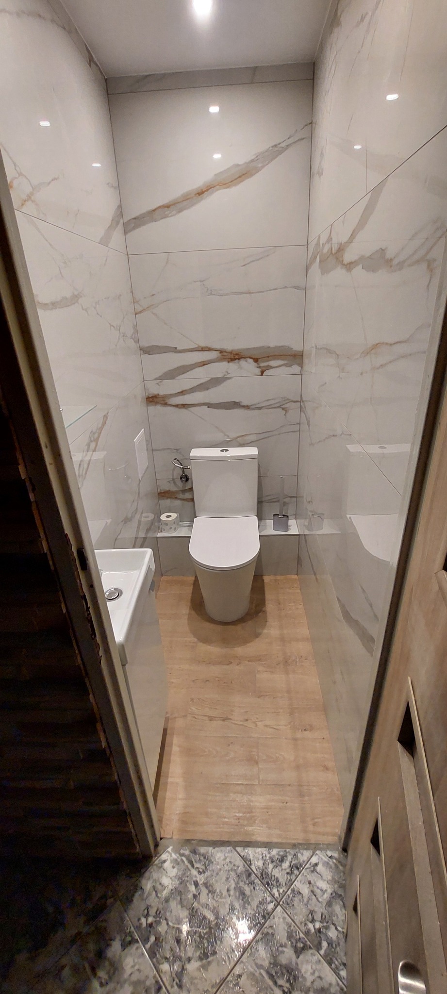 Wąska toaleta wykończona płytkami imitującymi marmur na ścianach i drewnem na podłodze, widok przez częściowo otwarte drewniane drzwi.