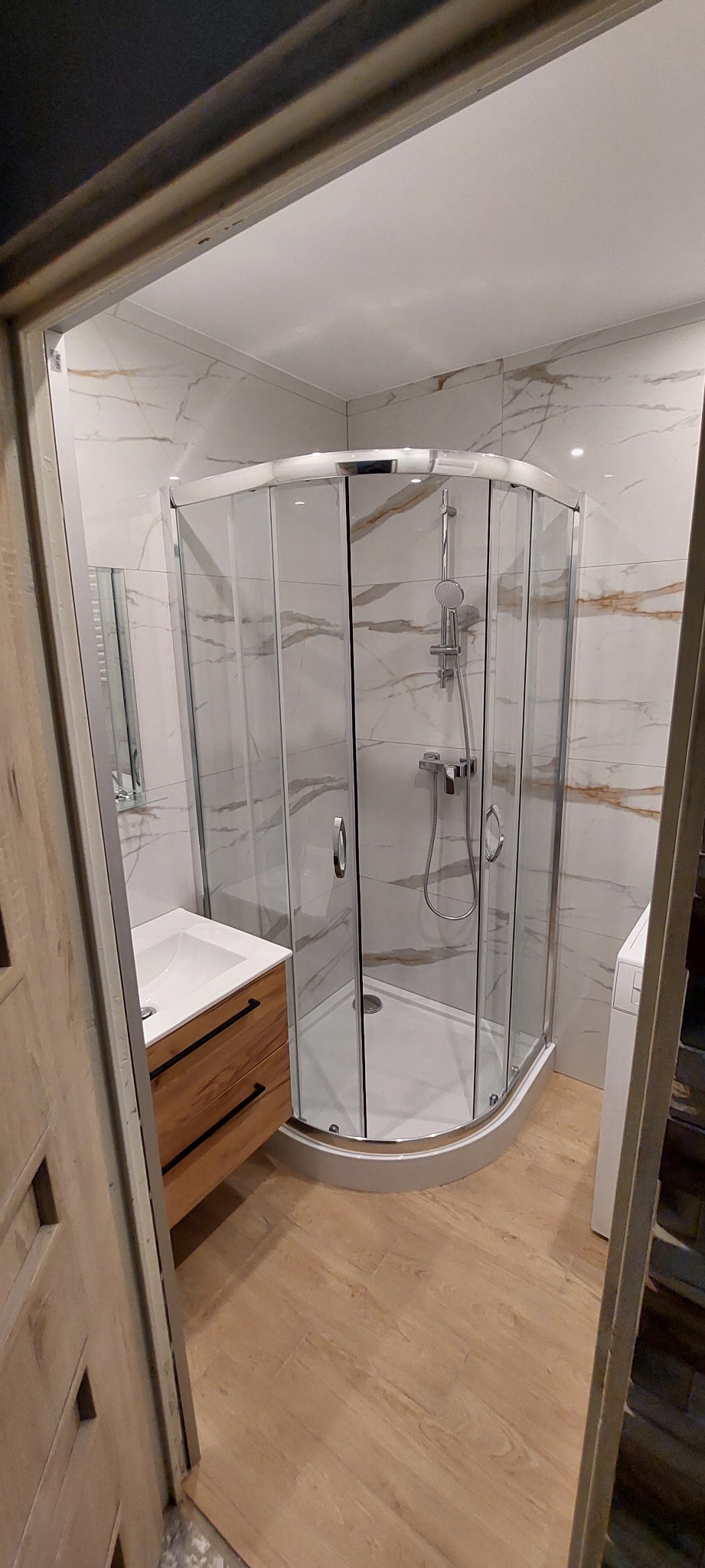 Nowoczesna łazienka z kabiną prysznicową narożną, umywalką w szafce z drewnianymi frontami i marmurowymi płytkami na ścianach, widok zza framugi drzwi.