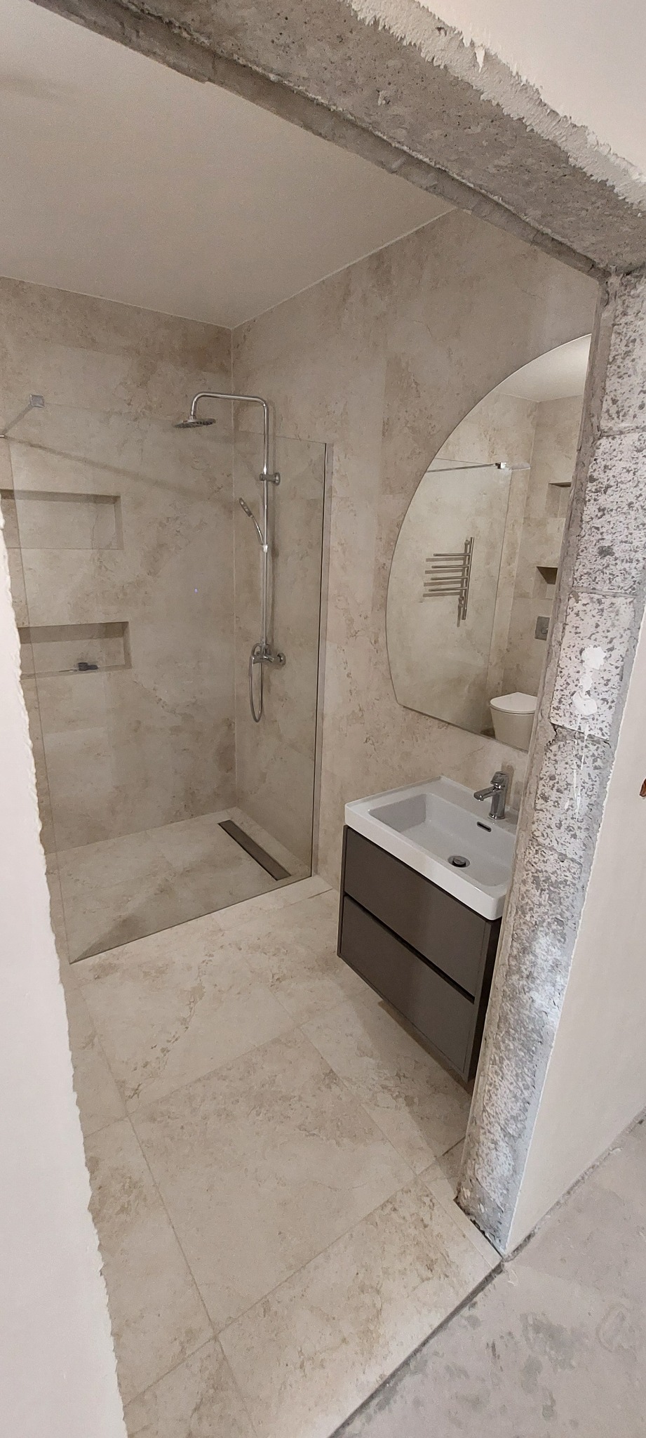 Nowoczesna łazienka w beżowych płytkach z kabiną prysznicową typu walk-in, umywalką z szafką i asymetrycznym lustrem, widoczny surowy betonowy fragment framugi drzwi.