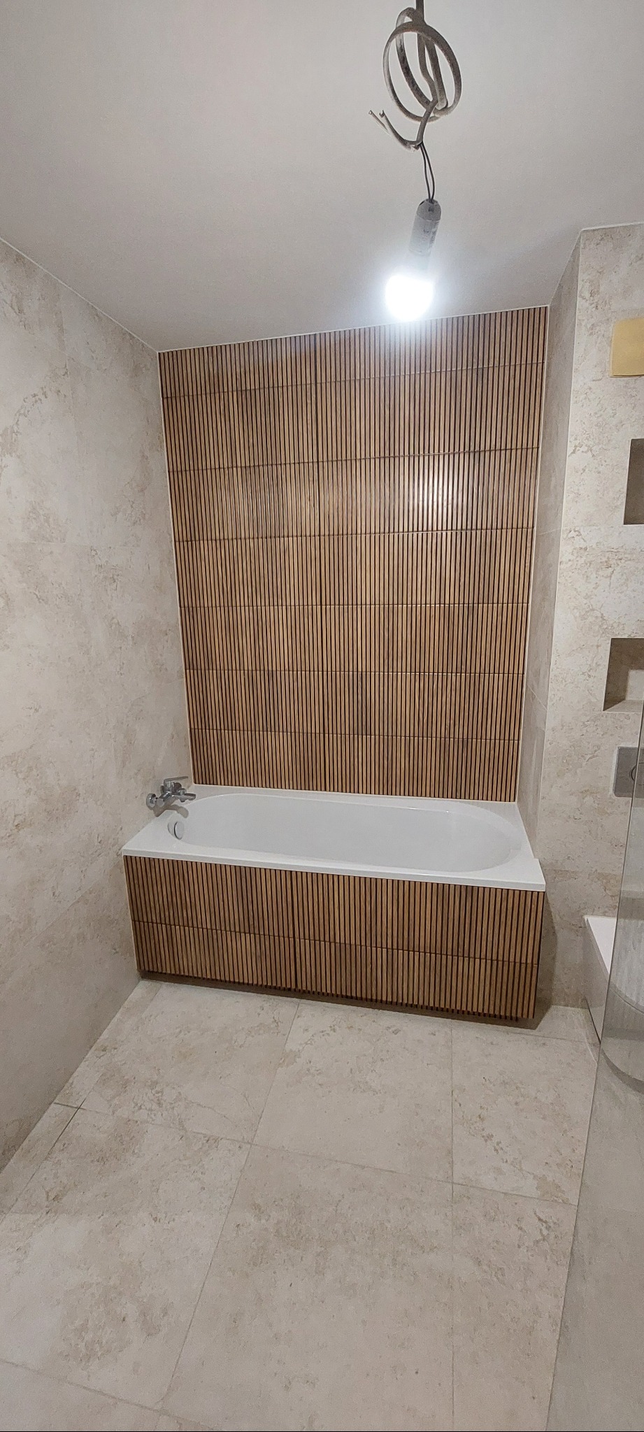 Nowoczesna łazienka z wanną obudowaną panelami drewnopodobnymi, ściana za wanną wykończona identycznymi panelami, kremowe płytki na podłodze i ścianach, widoczna niedokończona instalacja...