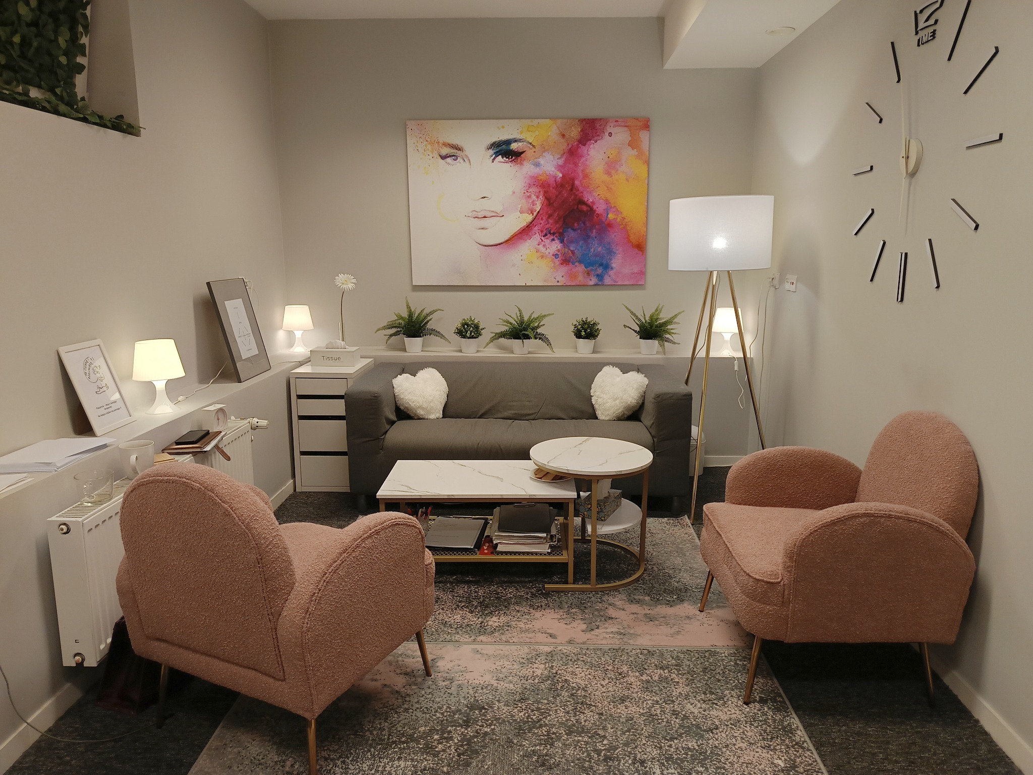 Jasne wnętrze gabinetu z dwoma różowymi fotelami, szarą sofą z poduszkami w kształcie serc, stolikiem kawowym, lampą podłogową i abstrakcyjnym obrazem na ścianie.