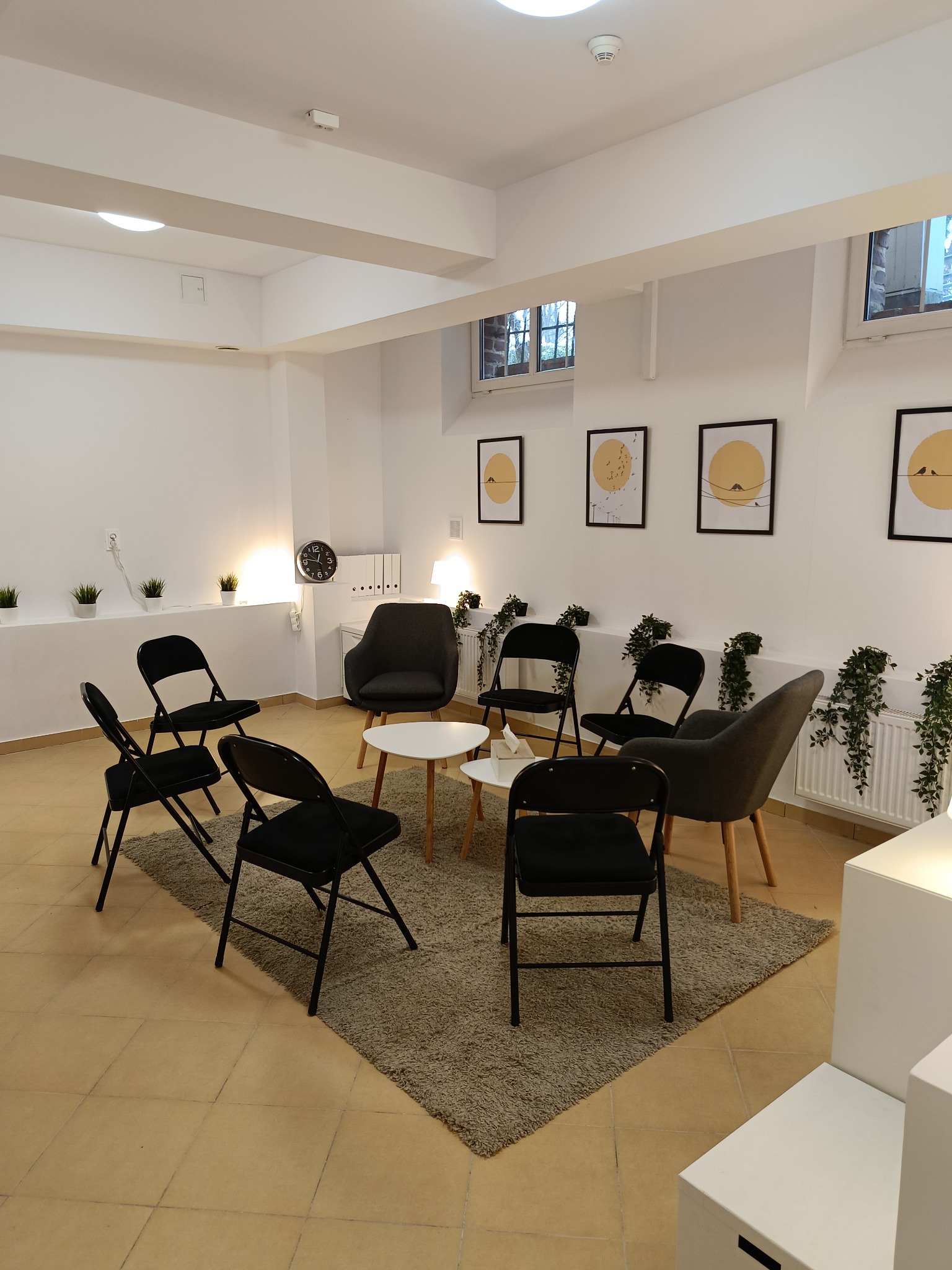 Jasne wnętrze sali terapeutycznej z krzesłami ustawionymi w kręgu na dywanie, minimalistyczne obrazy na ścianach, rośliny doniczkowe i zegar ścienny.