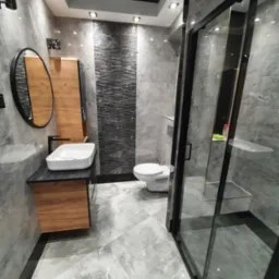 Nowoczesna łazienka w szarościach z drewnianymi akcentami, owalne lustro z czarną ramą, biała umywalka nablatowa na czarnym blacie, kabina prysznicowa typu walk-in z czarnymi profilami, toaleta...