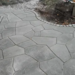 Ścieżka z szarej kostki o nieregularnym kształcie, obrzeża wykończone kostką granitową, widoczne fragmenty trawnika i ściana budynku.