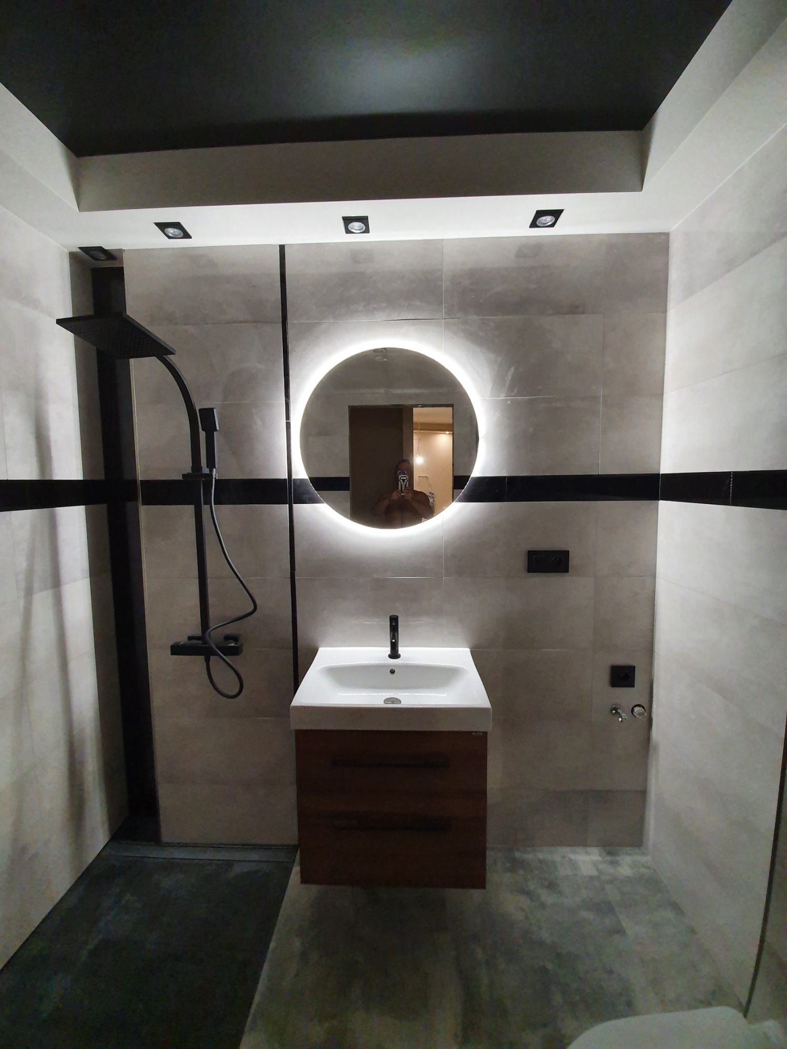 Nowoczesna łazienka z kabiną prysznicową typu walk-in, umywalką z szafką z drewnianym frontem i okrągłym lustrem podświetlanym LED, czarne akcenty armatury i płytek