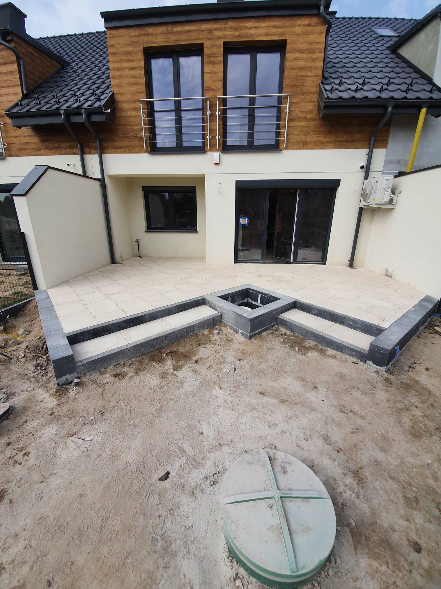 Taras z jasnych płyt, z dwoma stopniami obłożonymi szarą kostką, wbudowane palenisko, widoczne przygotowane podłoże i pokrywa studzienki kanalizacyjnej.