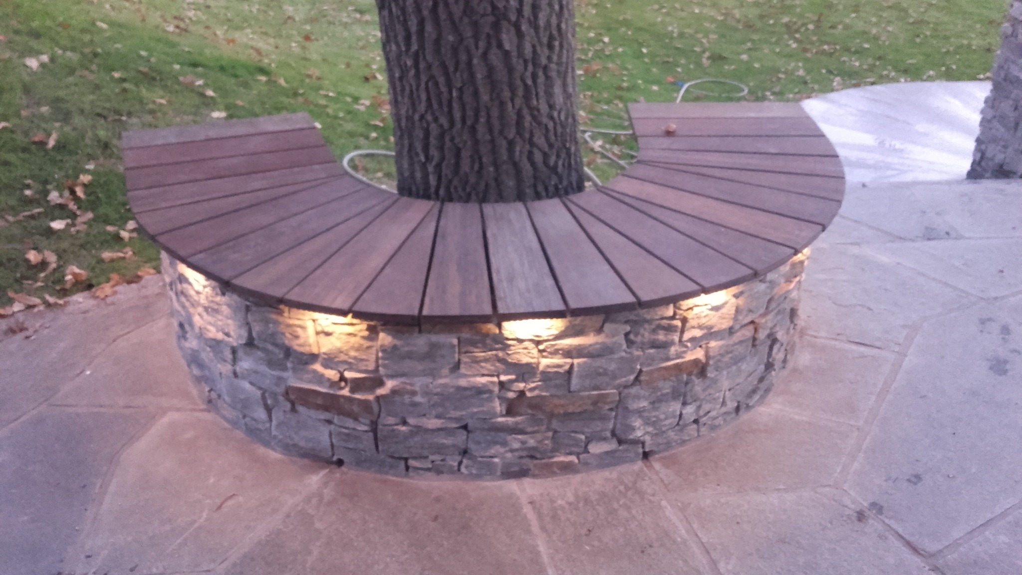 Półokrągła ławka z drewnianym siedziskiem otaczająca drzewo, podświetlona od dołu kamienną obudową z wbudowanymi lampkami punktowymi.