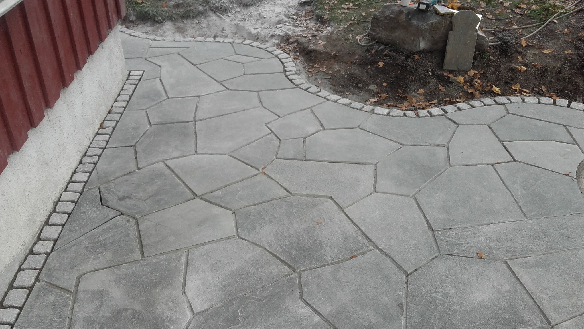 Ścieżka z szarej kostki o nieregularnym kształcie, obrzeża wykończone kostką granitową, widoczne fragmenty trawnika i ściana budynku.