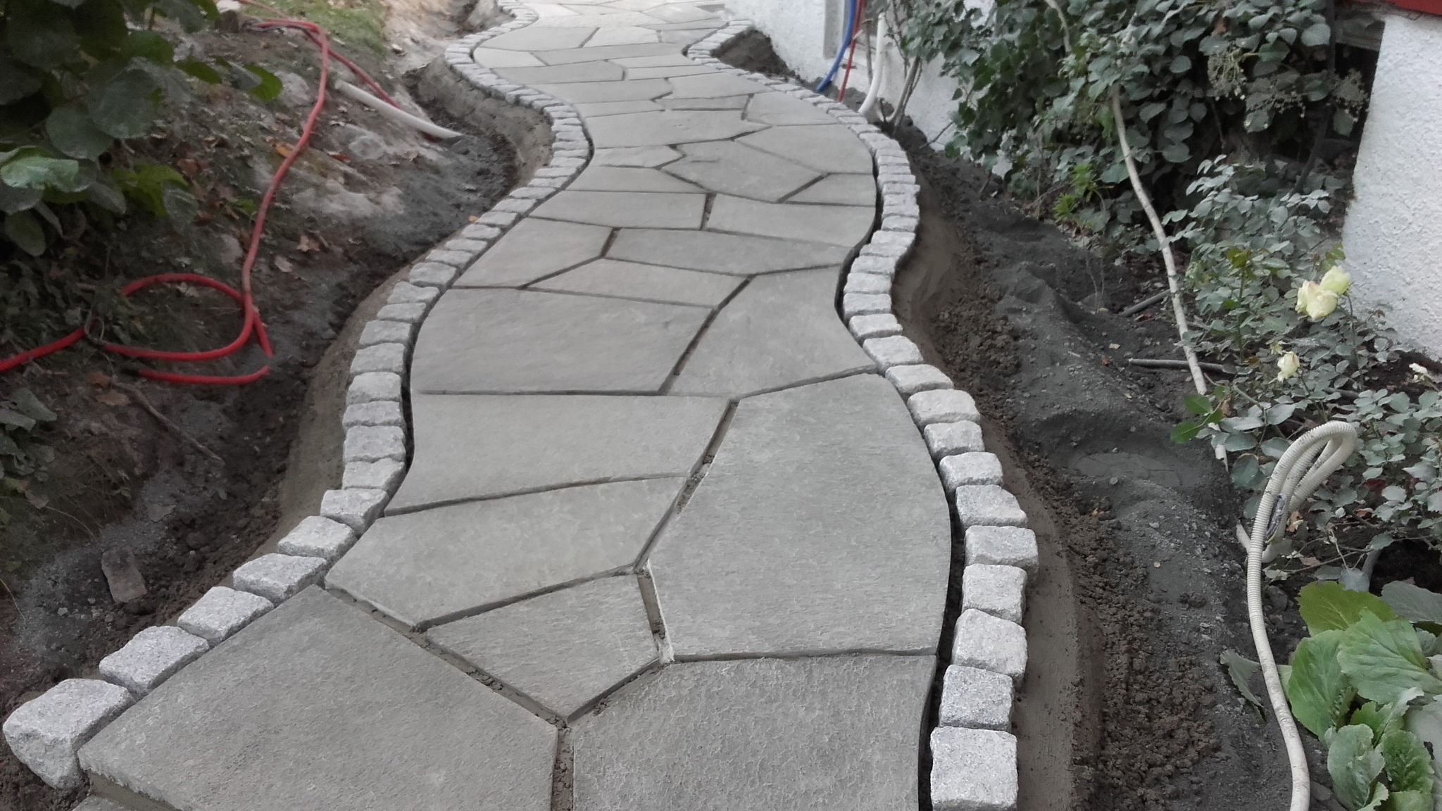 Kręta ścieżka z nieregularnych płyt kamiennych obramowana kostką granitową, prowadząca przez ogród.