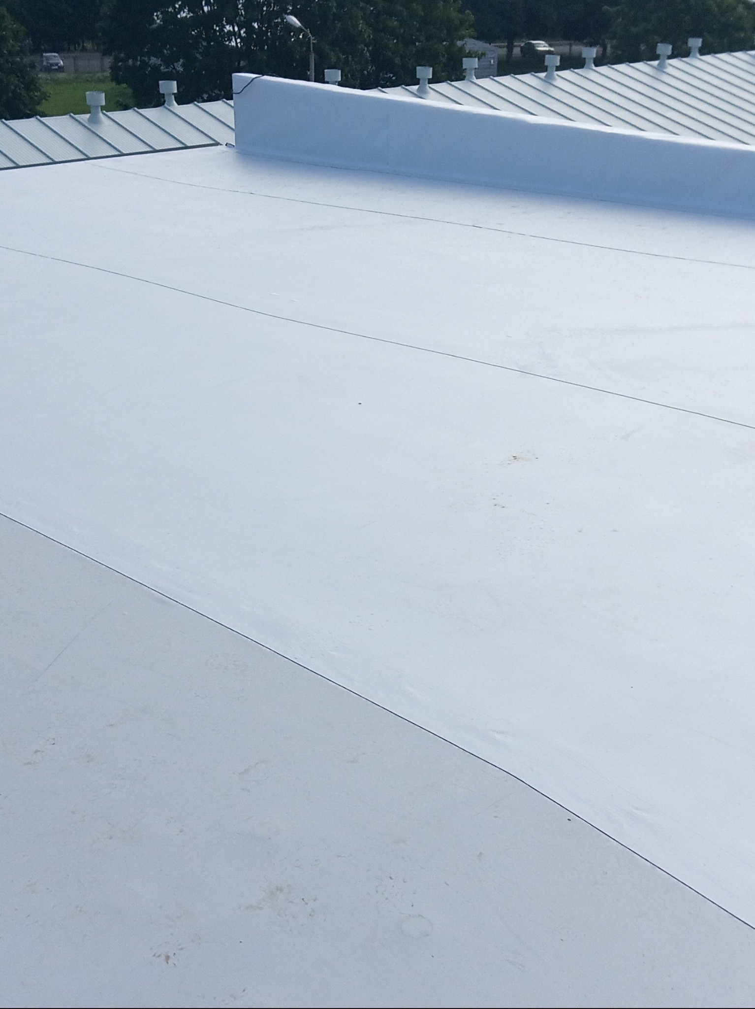 Biała, płaska powierzchnia dachu z widocznymi liniami łączeń, w tle fragment dachu blaszanego z kominami wentylacyjnymi i drzewami.