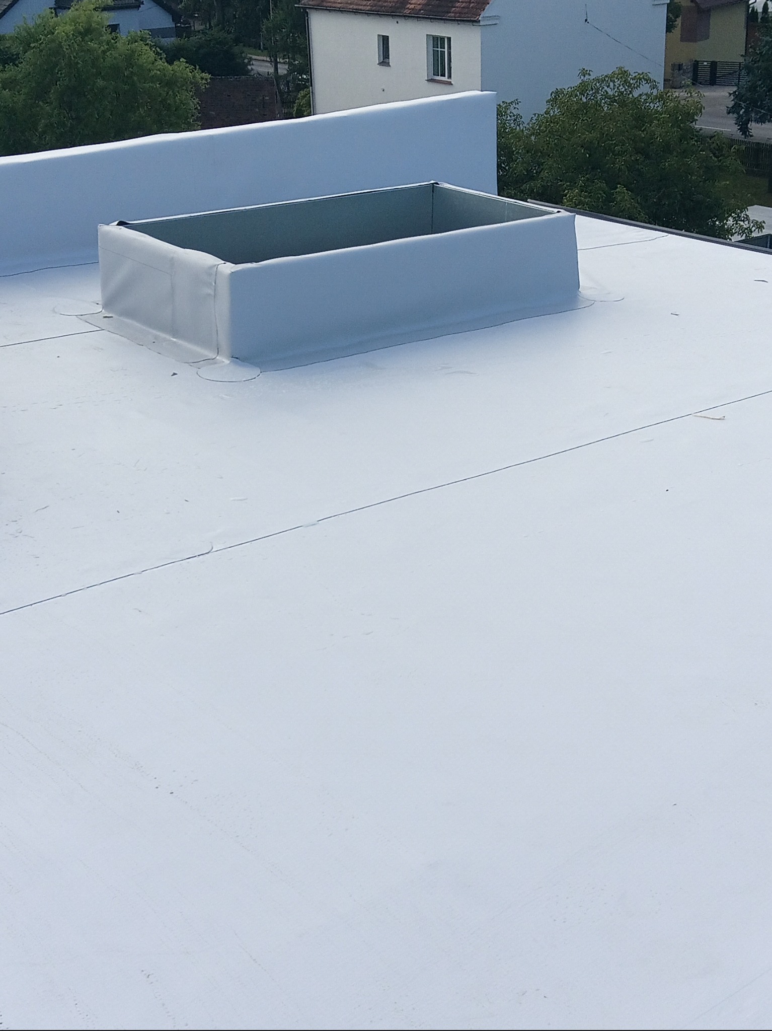 Jasny, płaski dach z obróbką blacharską komina wentylacyjnego. Widoczne budynki w tle. Minimalistyczny design z precyzyjnym wykończeniem.