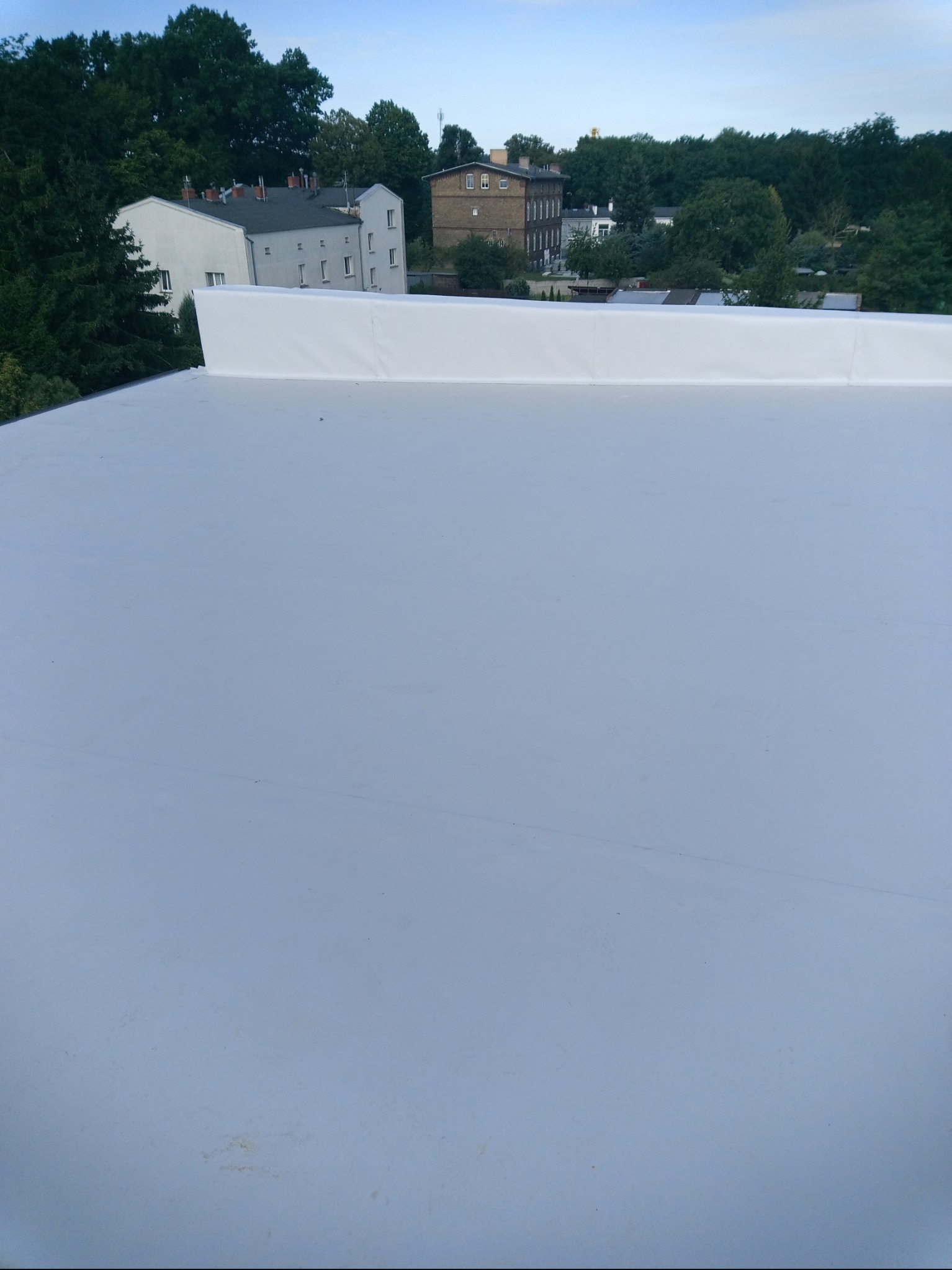 Biały, płaski dach z widocznym fragmentem attyki i zabudową na tle zieleni oraz zabudowań miejskich w oddali. Ujęcie z góry, prezentujące gładką powierzchnię.