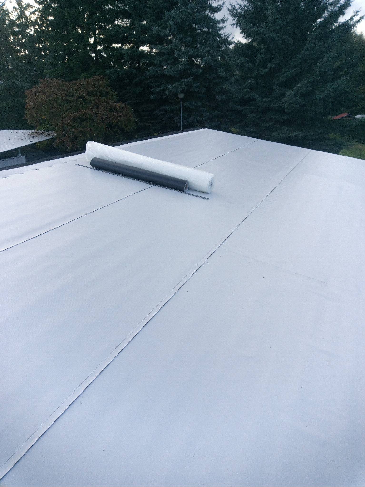 Szary dach z papy termozgrzewalnej, na którym leżą dwie rolki papy: biała i czarna. W tle drzewa iglaste i liściaste. Widok z góry.