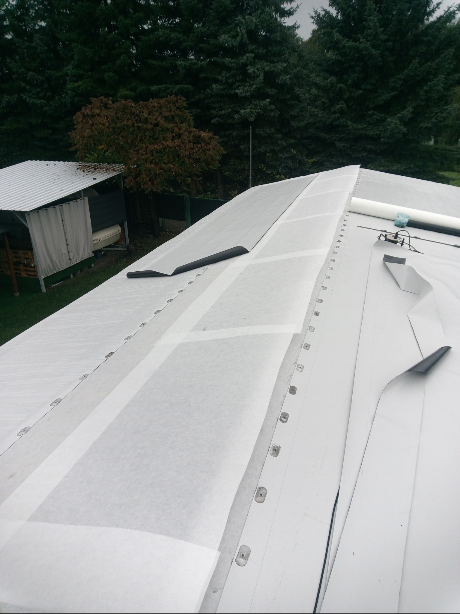 Biała membrana dachowa z widocznymi mocowaniami, częściowo zakryta, w tle zieleń drzew i fragment budynku z blachy falistej.