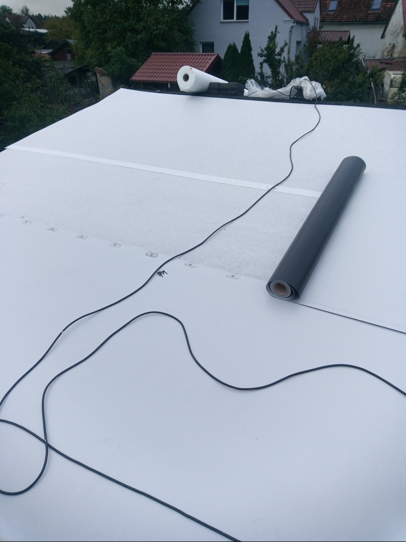 Biała membrana dachowa z rolką czarnej papy i przewodami na płaskim dachu. W tle zabudowa jednorodzinna i zieleń.