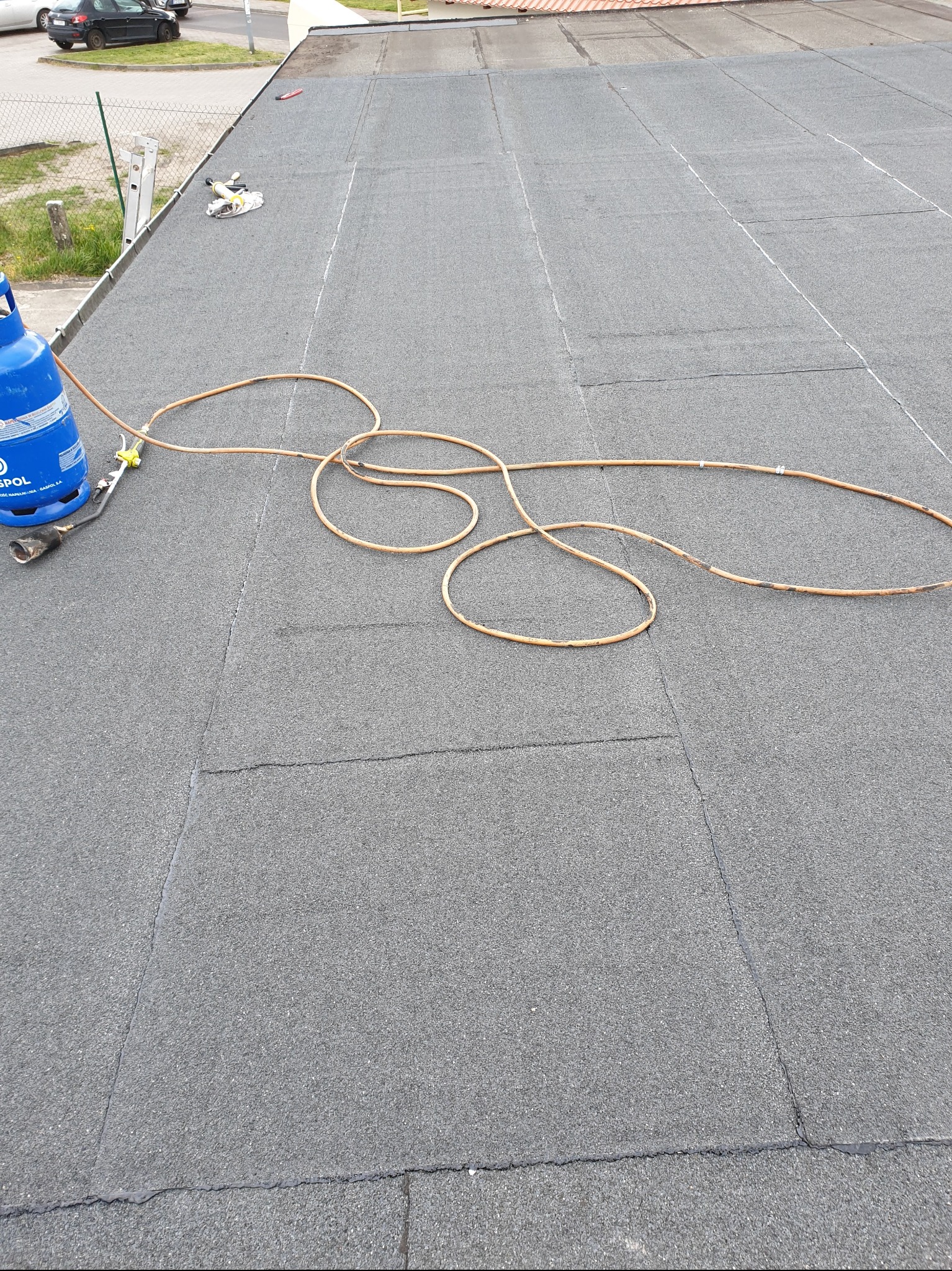 Szary, płaski dach pokryty papą termozgrzewalną z widocznym sprzętem dekarskim: butla gazowa, palnik i węże. Widoczne łączenia papy. Dach w trakcie prac.