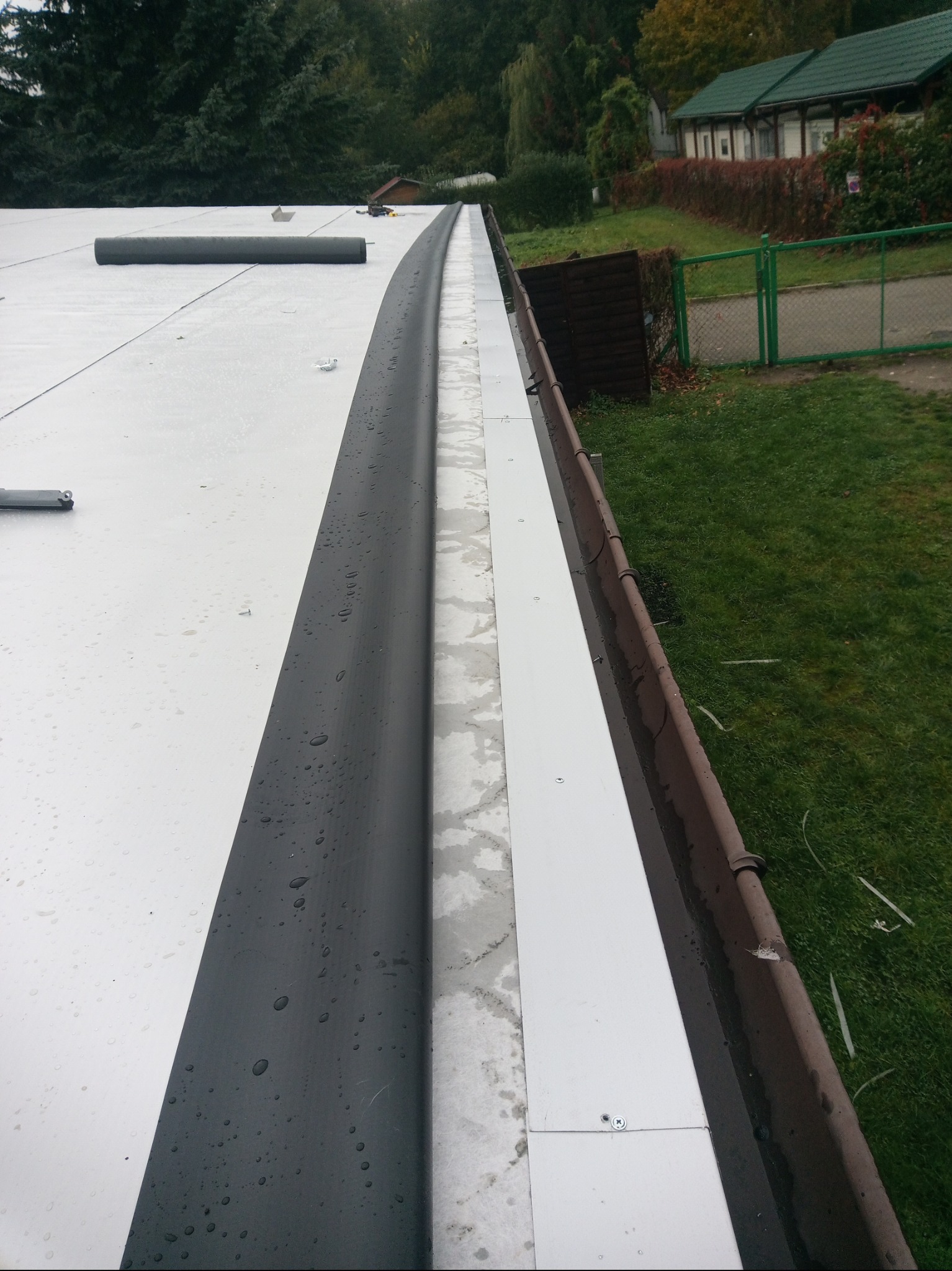 Widok z góry na płaski, biały dach z czarną obróbką blacharską i brązową rynną. W tle zieleń drzew i budynki.