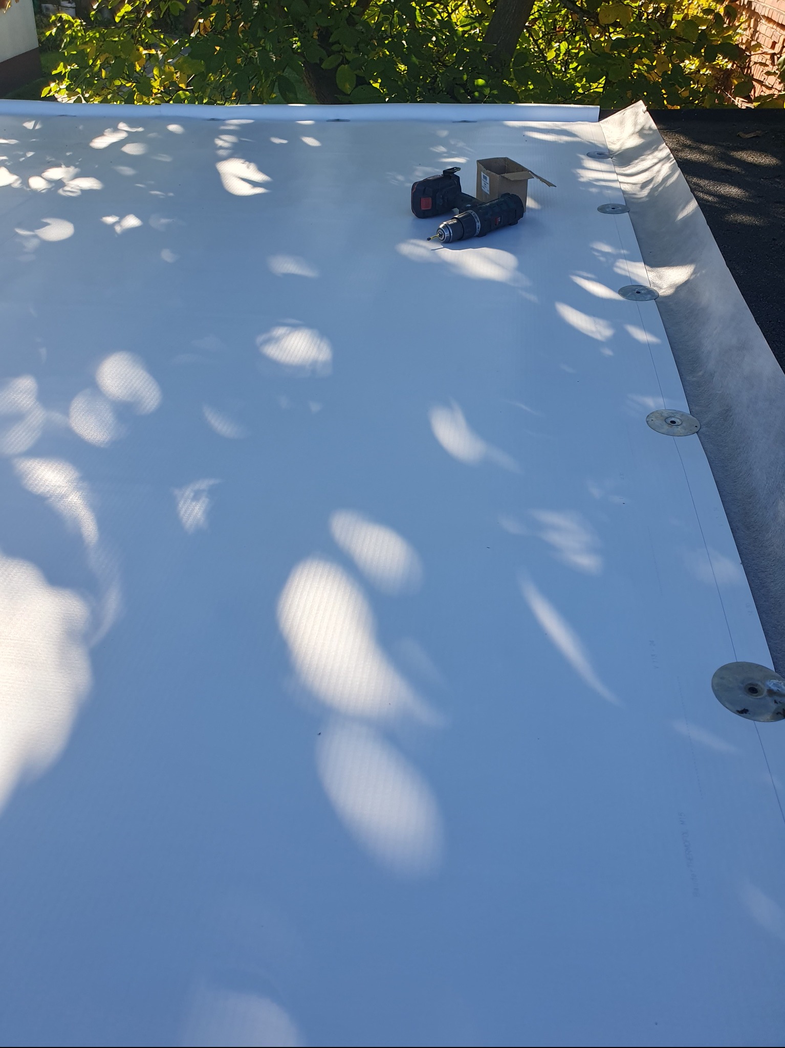 Biała membrana dachowa z widocznymi mocowaniami, narzędzia pracy (wiertarka, miarka) i cień drzewa. Dach płaski w trakcie prac dekarskich, widok z góry.