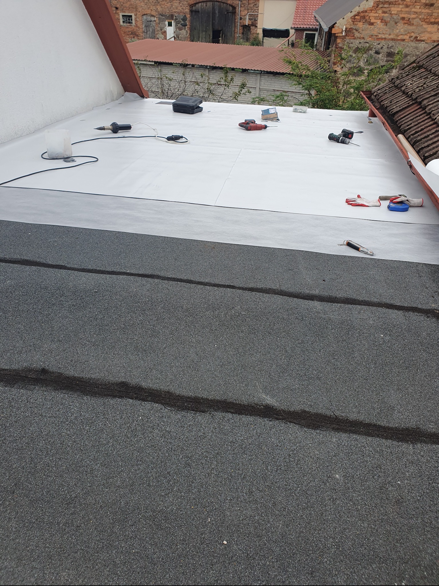 Dach pokryty papą termozgrzewalną i membraną PVC, widoczne narzędzia dekarskie i rękawice robocze. Tło stanowi budynek z czerwonej cegły.
