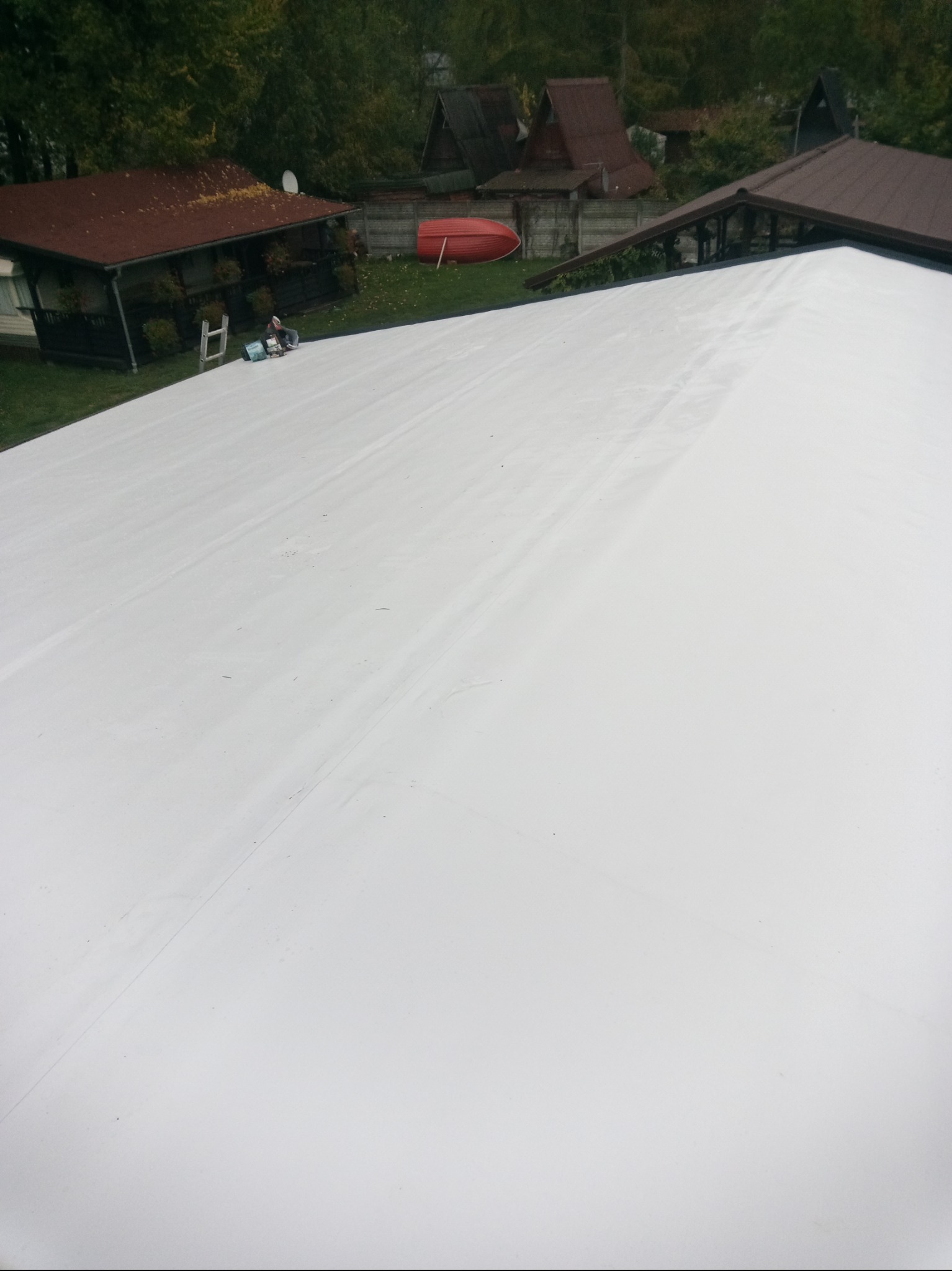 Biała, gładka powierzchnia dachu po kryciu, z widocznym fragmentem ogrodu i zabudowań w tle, w tym czerwona łódka. Praca dekarska w toku.