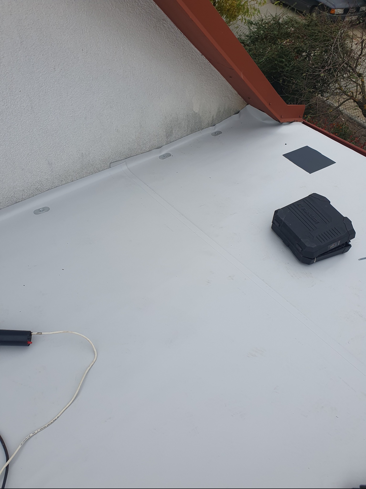 Szara membrana dachowa z elementami mocującymi, narzędziami i fragmentem obróbki blacharskiej w kolorze ceglastym. Widok z góry, częściowo widoczny budynek i zieleń.