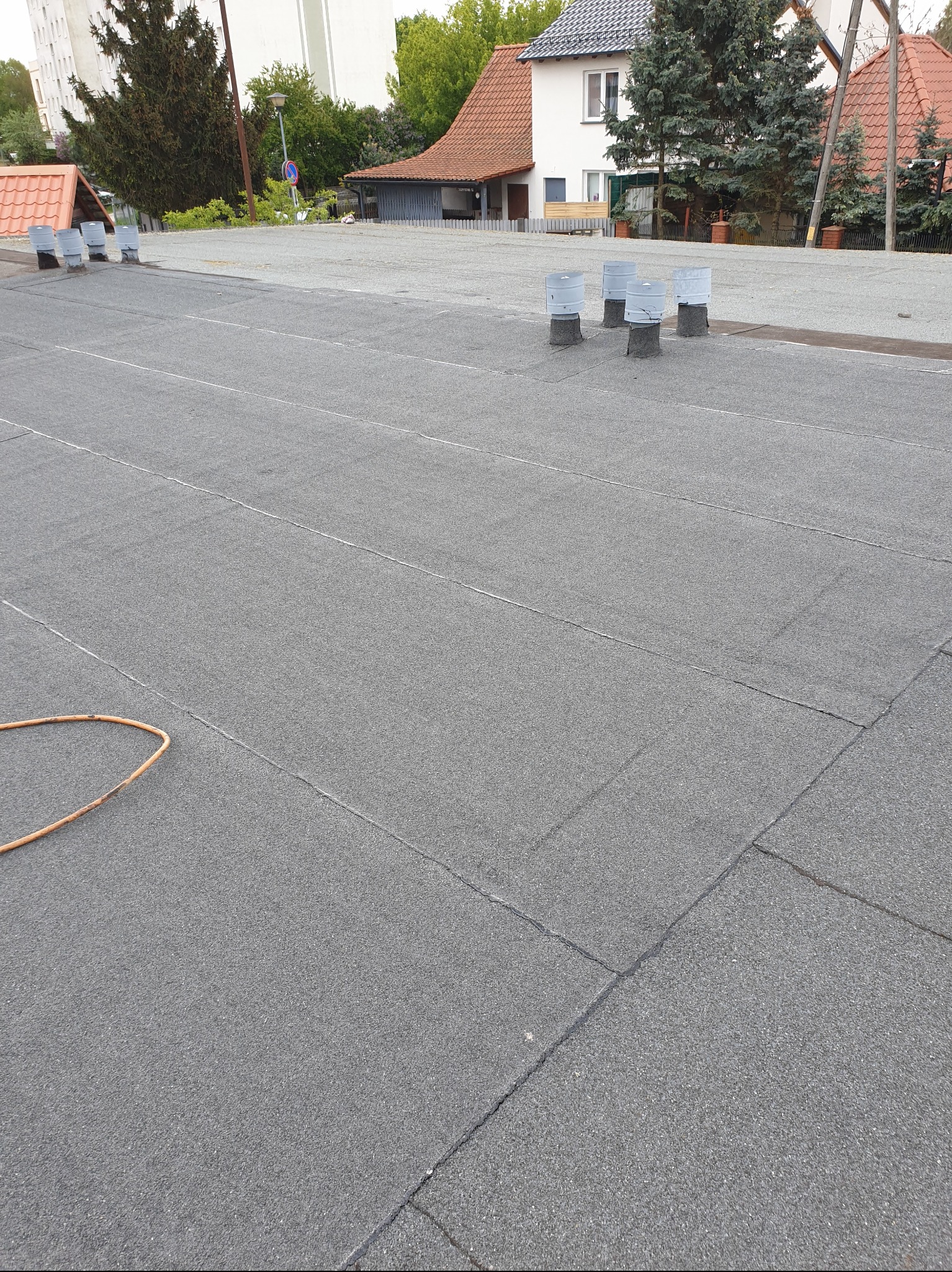 Szary dach pokryty papą bitumiczną, widoczne kominki wentylacyjne i fragment węża. Tło stanowią budynki i drzewa. Układ połaci dachowej z wyraźnymi łączeniami.