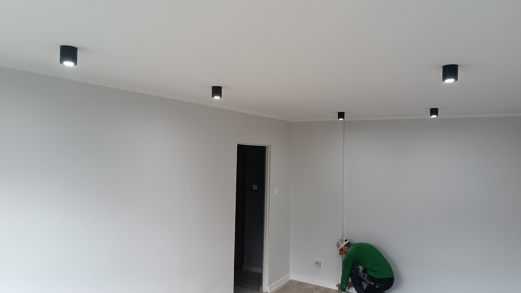 Pracownik w zielonej bluzie przy podłodze, wykańcza szpachlowaniem narożnik w świeżo pomalowanym na szaro pomieszczeniu z zamontowanymi czarnymi lampami sufitowymi.