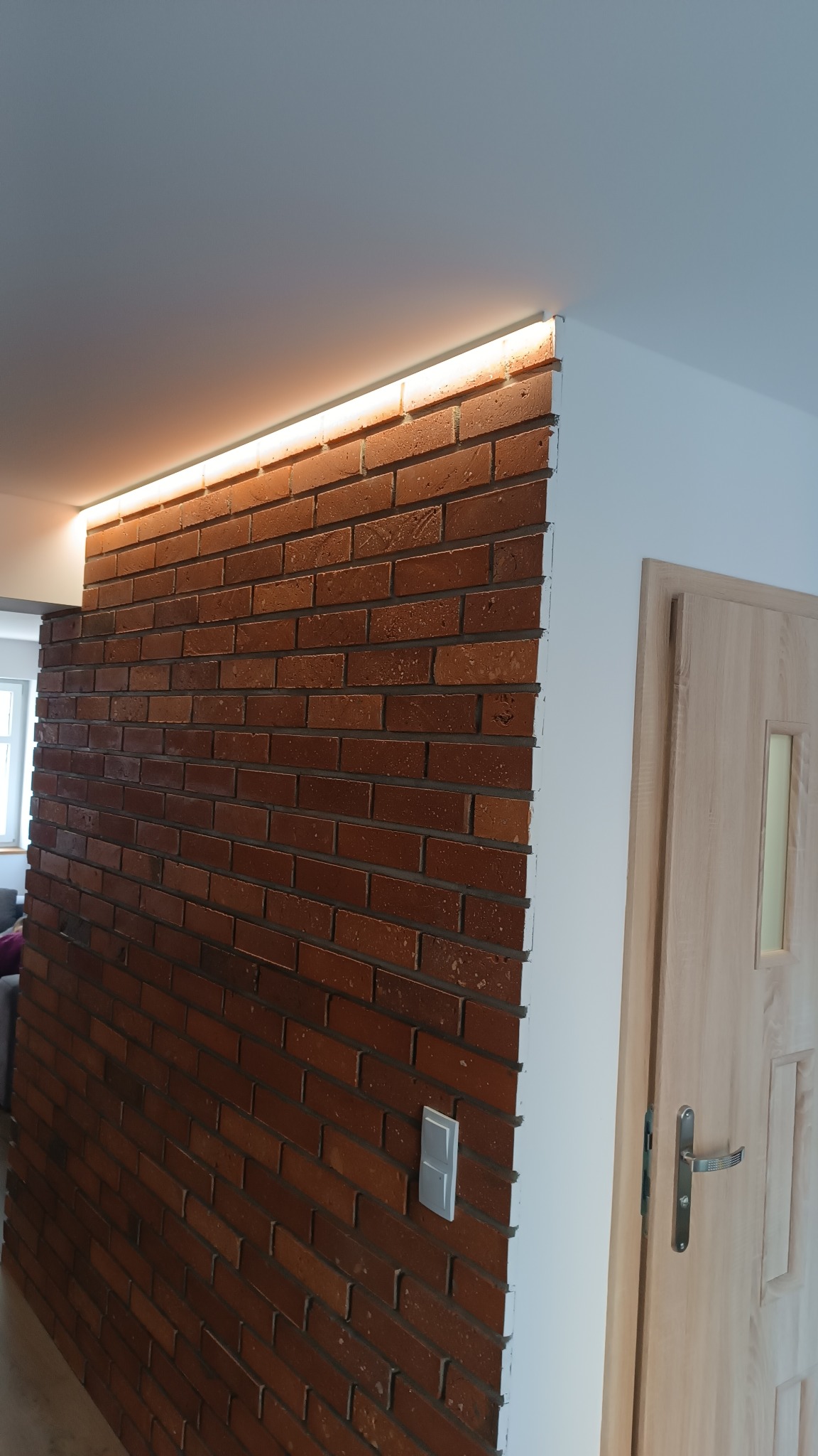 Ściana z cegły klinkierowej z ukrytym oświetleniem LED przy suficie, sąsiadująca z białą ścianą i drewnianymi drzwiami z przeszkleniem.