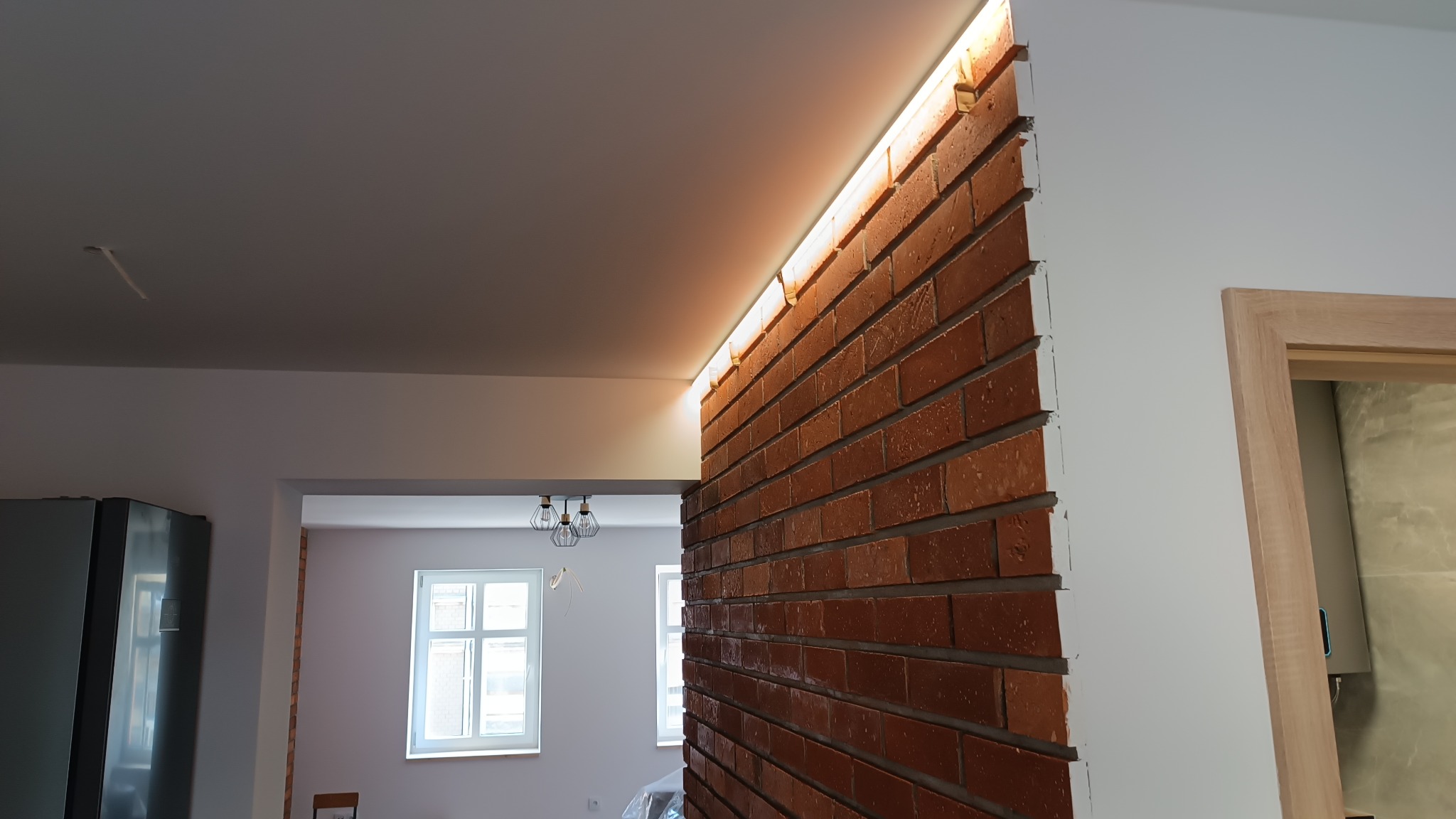 Ceglana ściana z podświetleniem LED w trakcie prac wykończeniowych, widoczny fragment kuchni i łazienki z surową ościeżnicą.