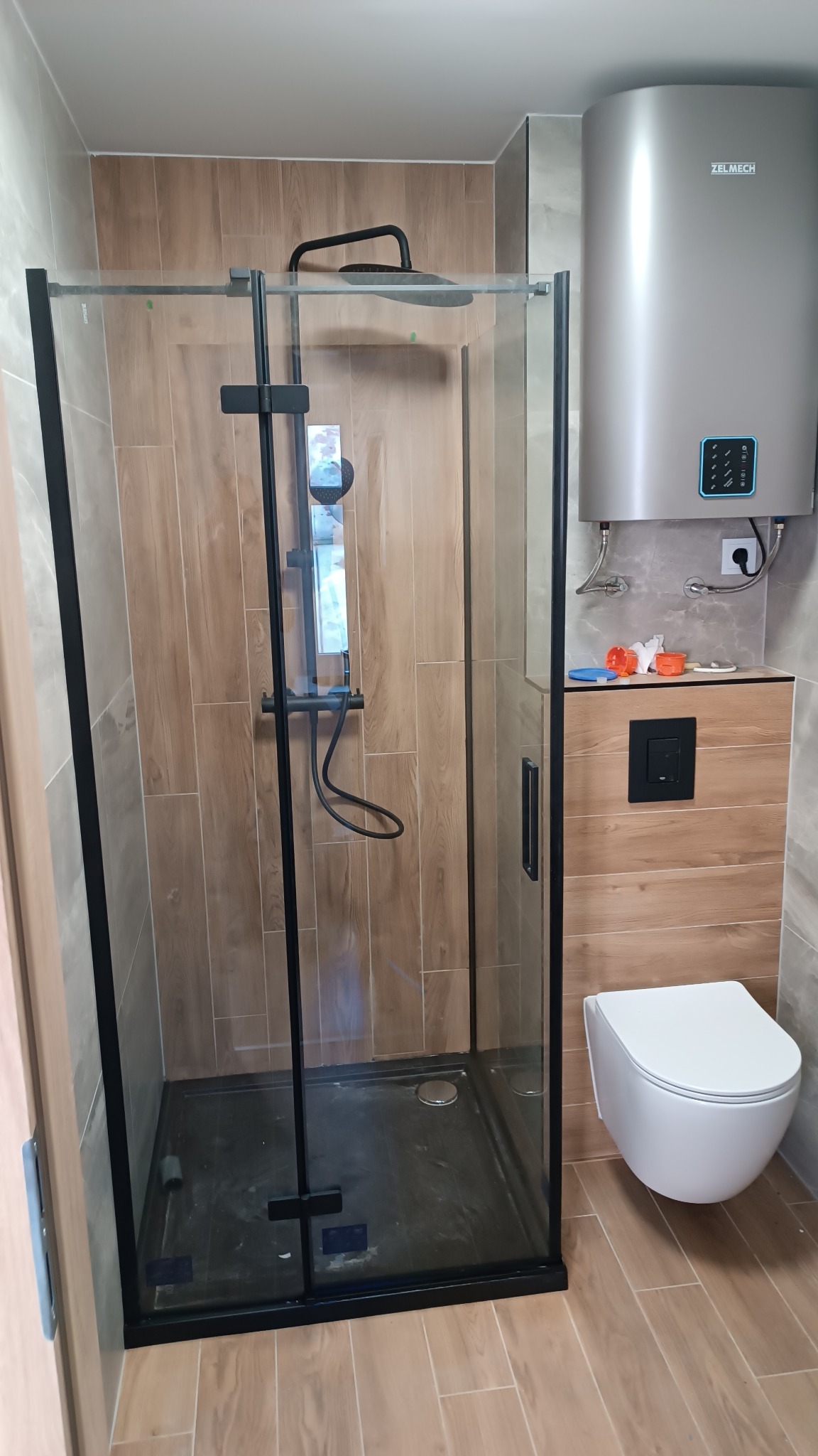 Kabina prysznicowa z czarnymi profilami i deszczownicą, obok podwieszana toaleta i bojler ZELMECH na ścianie wyłożonej płytkami imitującymi drewno.