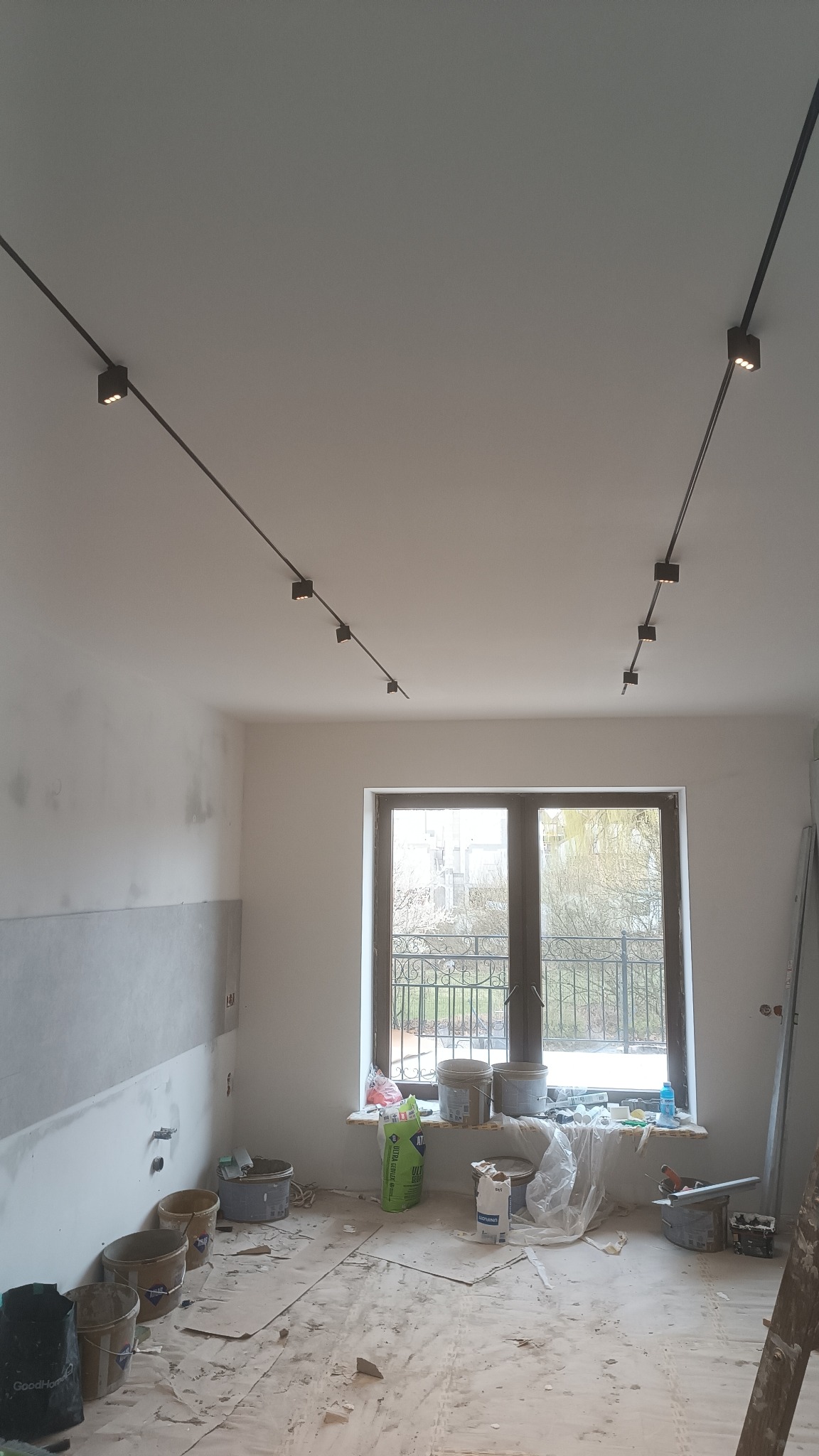 Pokój w trakcie prac wykończeniowych: ściany częściowo wyszpachlowane, nowe oświetlenie szynowe na suficie, widoczne wiadra z zaprawą i resztki materiałów na podłodze przy oknie.