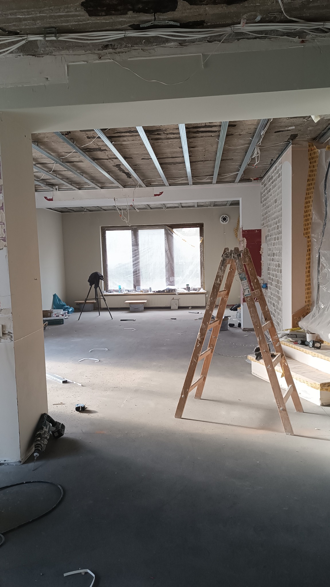 Wnętrze w trakcie prac: widoczne metalowe profile podwieszanego sufitu, drabina, wiertarka przy ścianie, okno z folią ochronną, ślady po kuciu na ścianie.