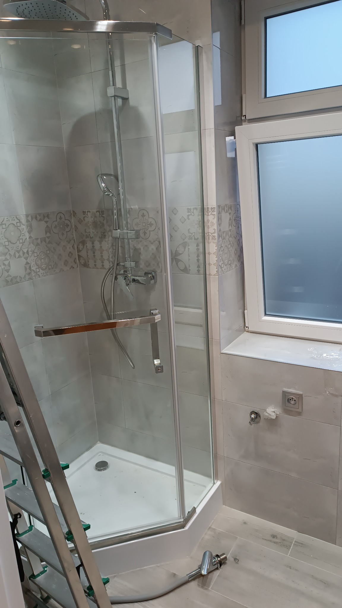 Nowa kabina prysznicowa z chromowanymi elementami i szklanymi drzwiami, zamontowana w łazience wykończonej jasnymi, wzorzystymi płytkami ceramicznymi, z widoczną drabinką i podłączeniem wodnym...