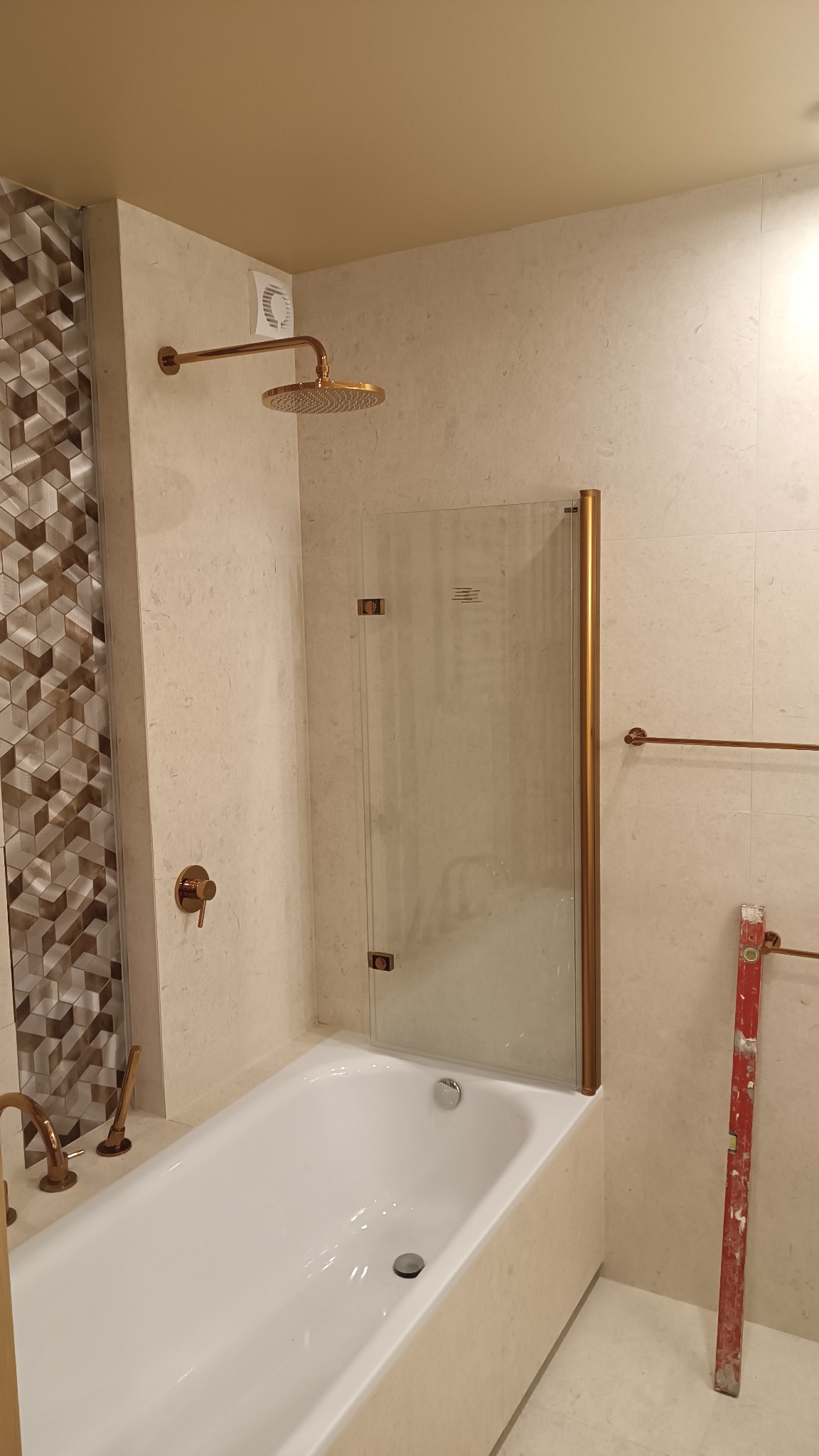 Nowoczesna łazienka z białą wanną, szklanym parawanem nawannowym z miedzianymi okuciami, deszczownicą w kolorze miedzi i dekoracyjną mozaiką na ścianie. Poziomica oparta o ścianę sugeruje trwające...