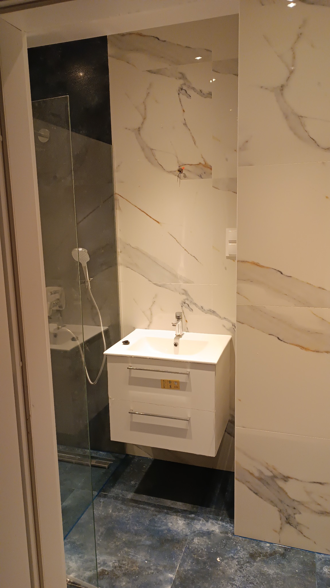 Nowoczesna łazienka z kabiną prysznicową ze szkła, białą szafką pod umywalkę z dwoma szufladami i marmurowymi płytkami na ścianach.