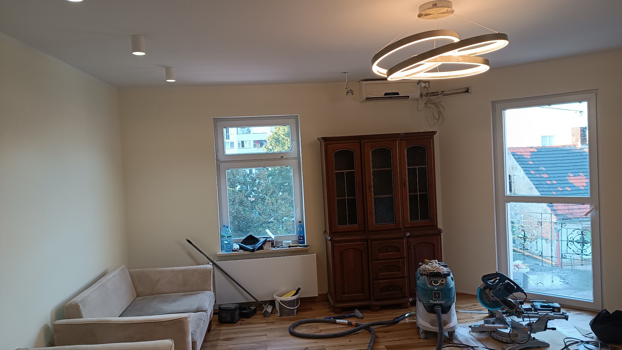 Pokój po remoncie z nową podłogą z paneli, widoczny sprzęt budowlany, drewniany kredens, sofa i nowoczesna lampa sufitowa.