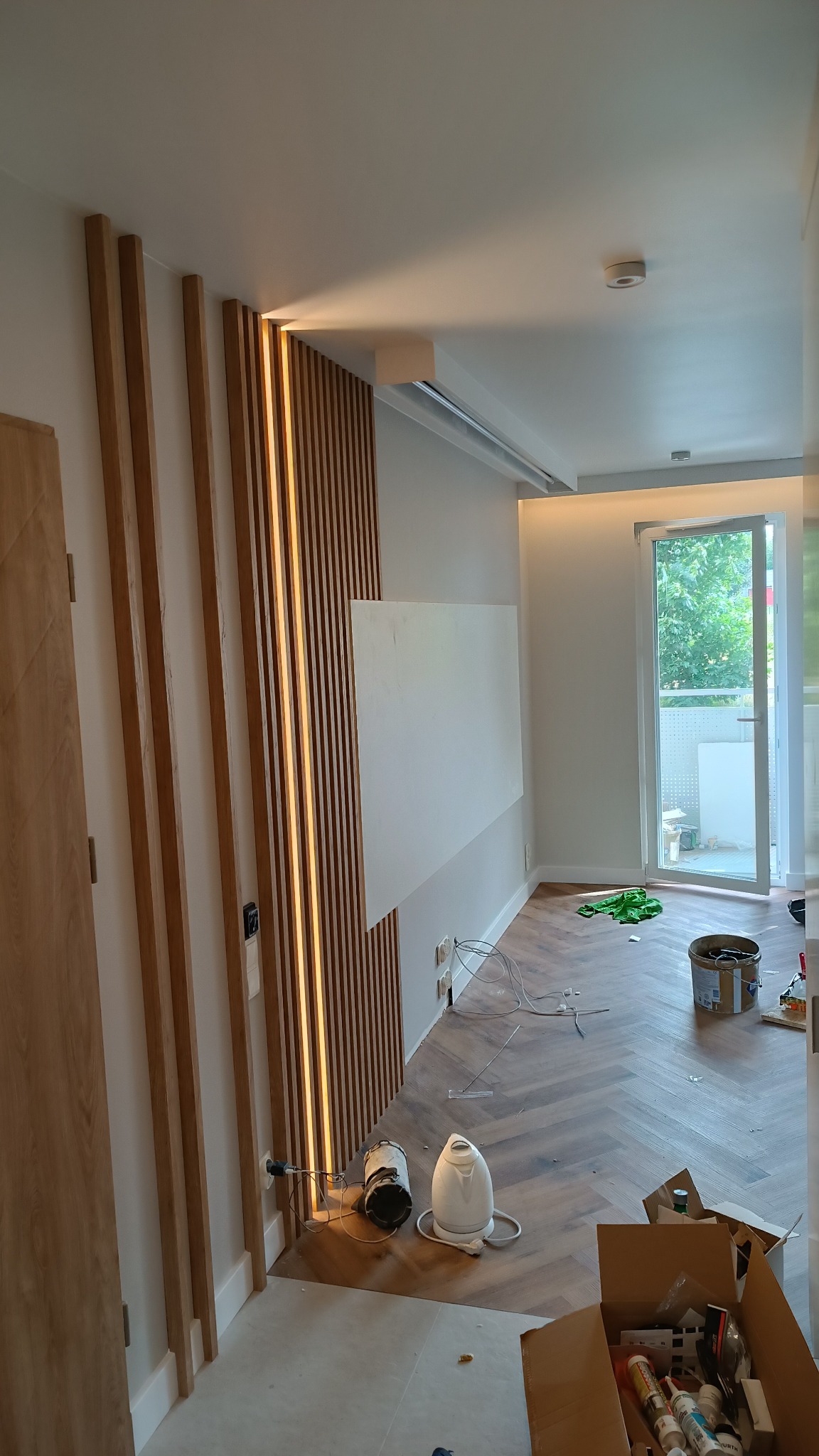Wnętrze w trakcie prac wykończeniowych: drewniane lamele dekoracyjne z oświetleniem LED na ścianie, częściowo zagruntowana ściana, parkiet ułożony w jodełkę, widoczne przewody elektryczne, karton...