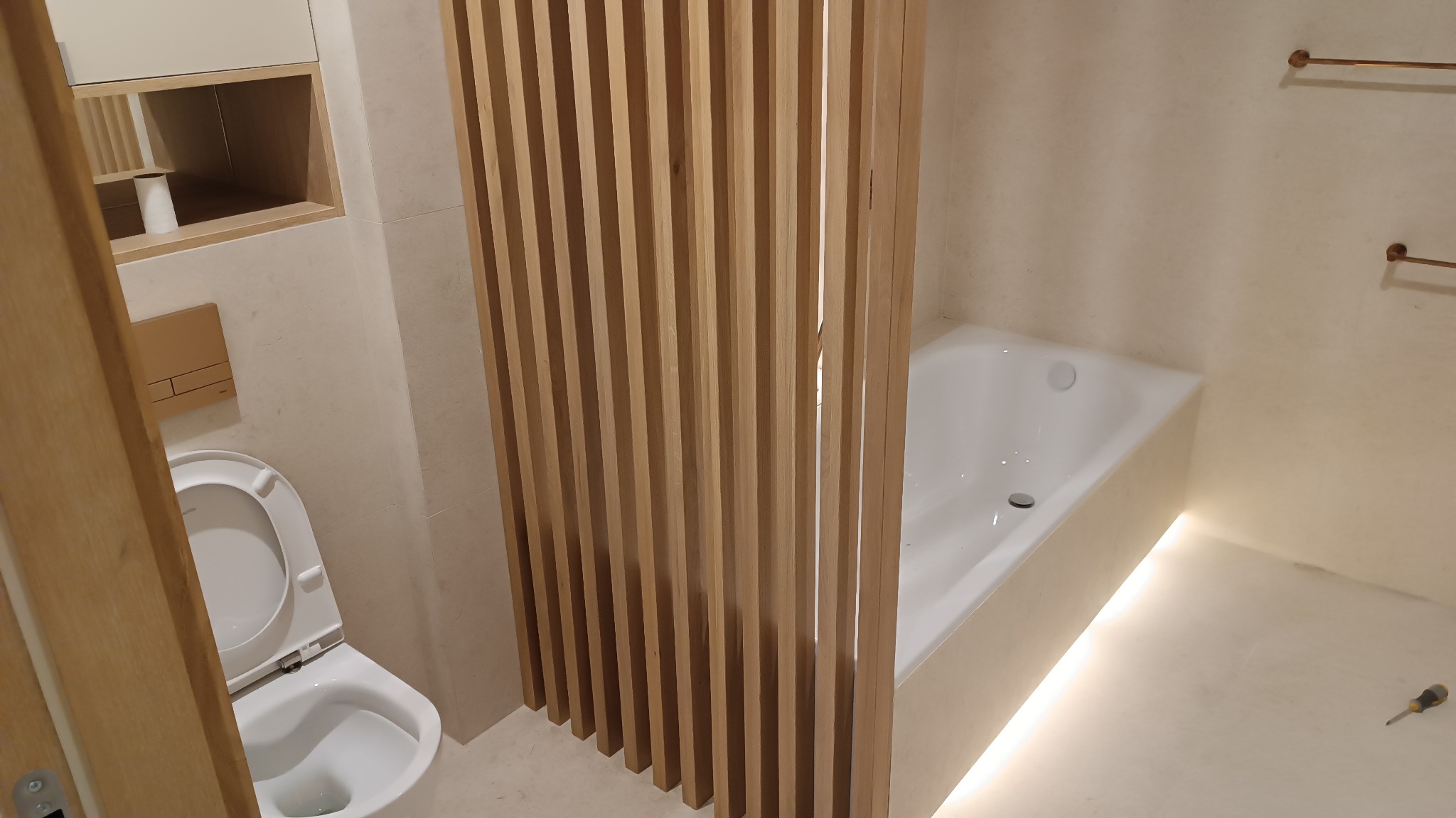 Nowoczesna łazienka z drewnianą ścianką działową, białą wanną podświetloną od dołu, wiszącą toaletą i minimalistycznymi wieszakami na ręczniki w kolorze miedzi.