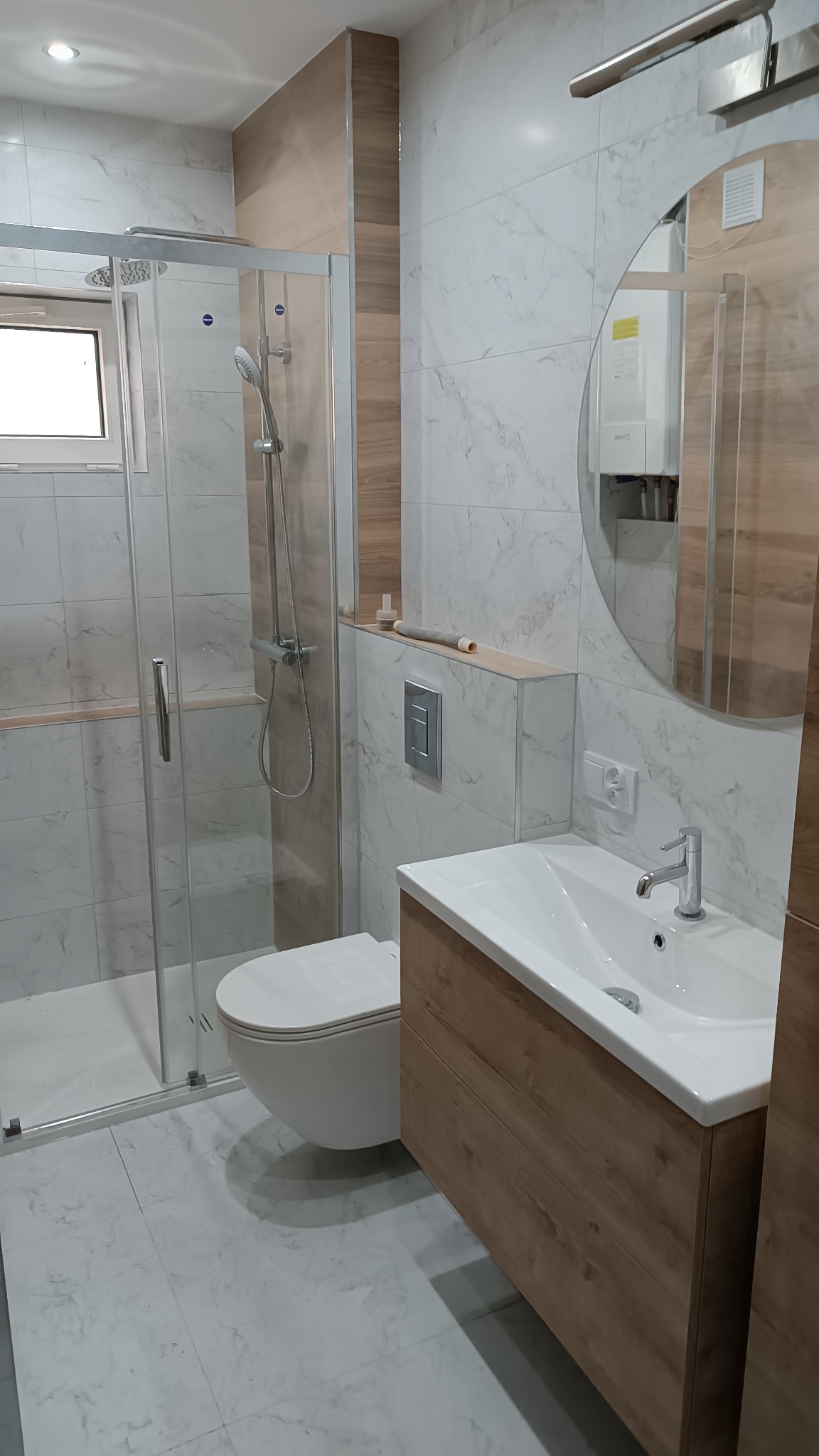 Nowoczesna łazienka z kabiną prysznicową, umywalką z szafką w odcieniach drewna i marmurowymi płytkami na ścianach i podłodze. Widoczny okrągłe lustro odbijające bojler. Na półce nad toaletą leżą...